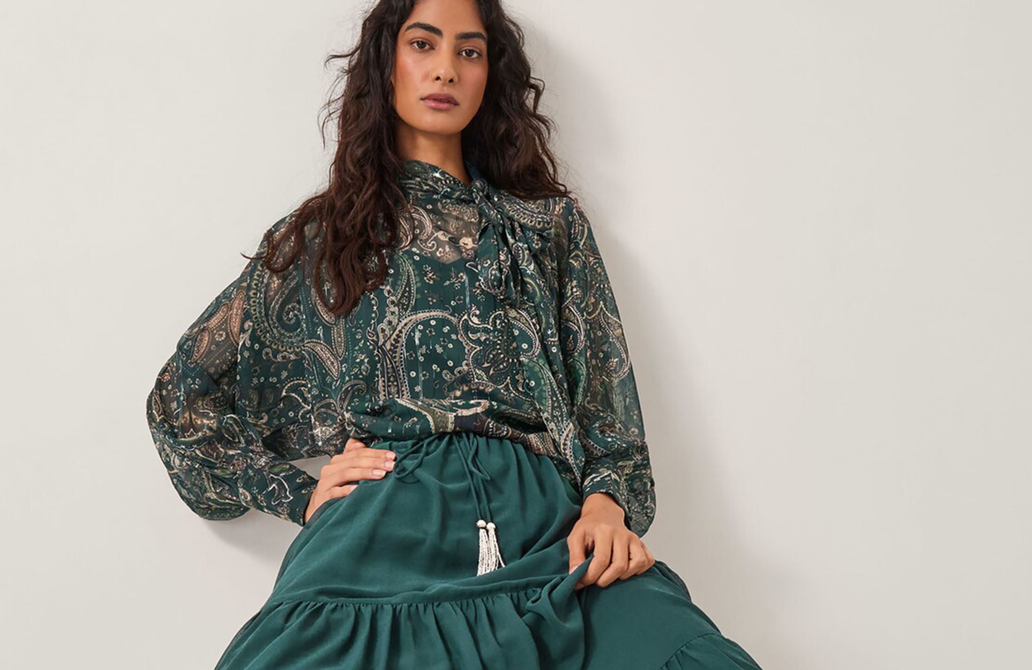 Model is wearing a green printed Monsoon Marianna sheer pussybow blouse with a teal Monsoon Holly tiered chiffon maxi skirt.