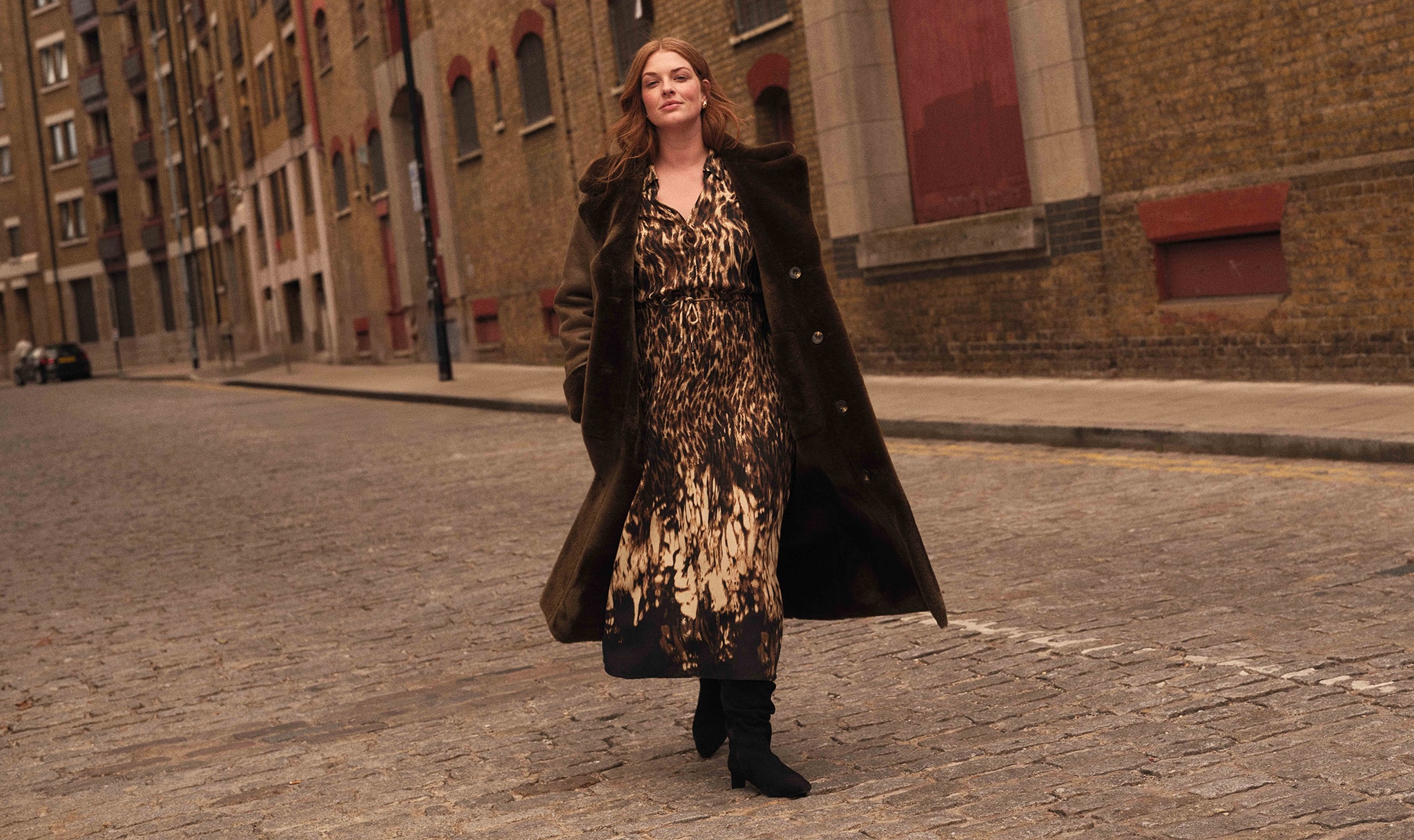 A lady walking in a cobbled street wearing a black and gold animal print dress with a think brown coat.