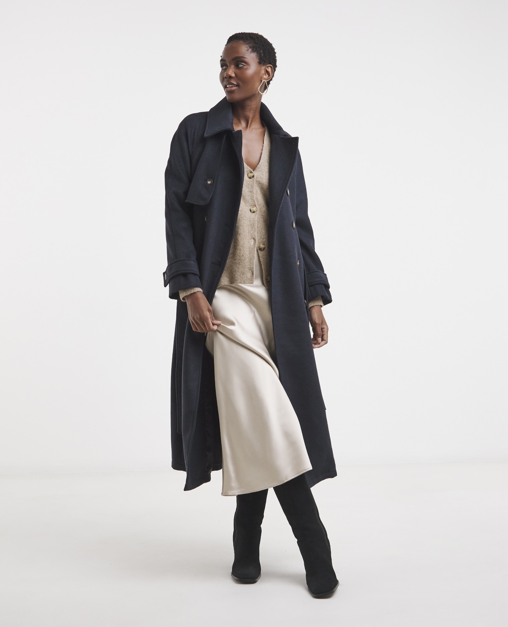 A woman wearing a long navy blue wool trench coat, a light gold cardigan, and a champagne-colored satin slip skirt, with black knee-high boots