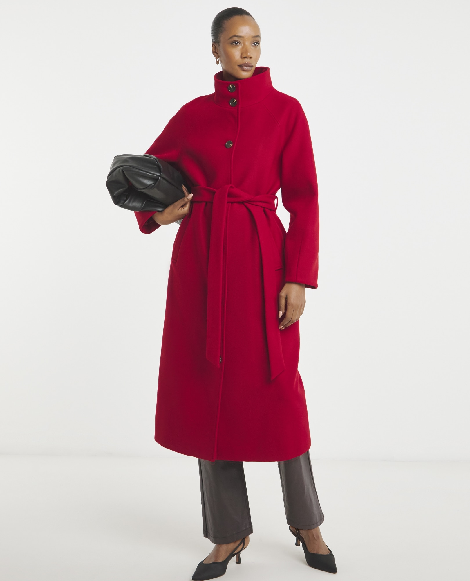 A woman standing in a long, bright red, high-neck coat with a tie belt, holding a black clutch bag, wearing black heels
