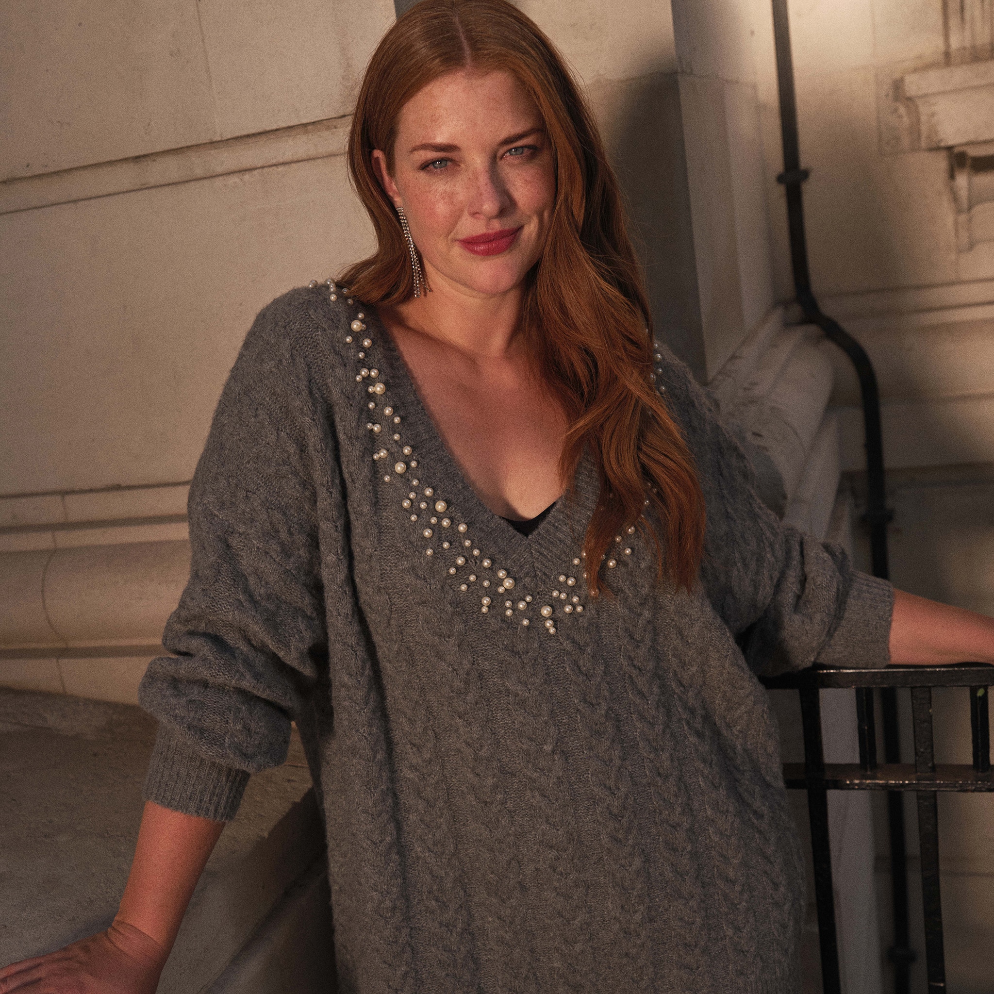 A lady wearing a grey v neck jumper with pearl style beads following the neck line.