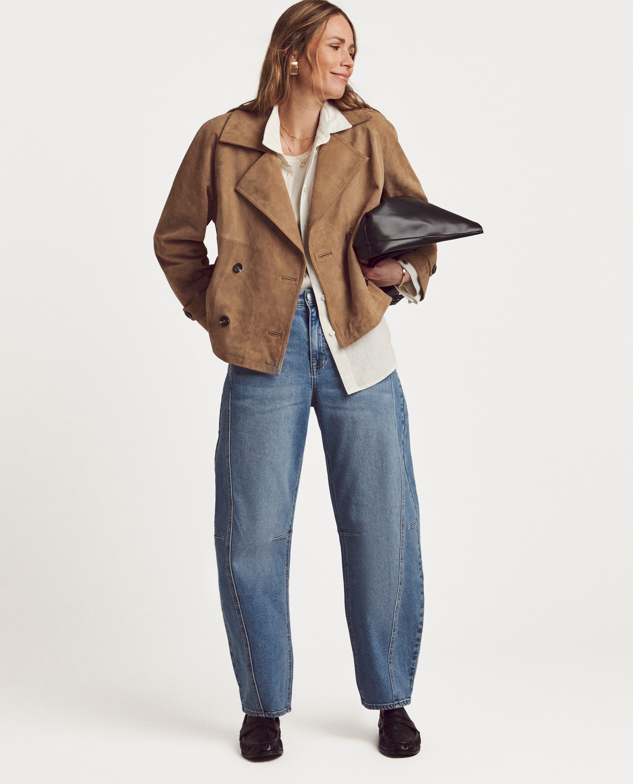 Model is wearing a sand suede short trench jacket with cream blouse and a pair of blue authentic tapered jeans with black loafers and handbag.