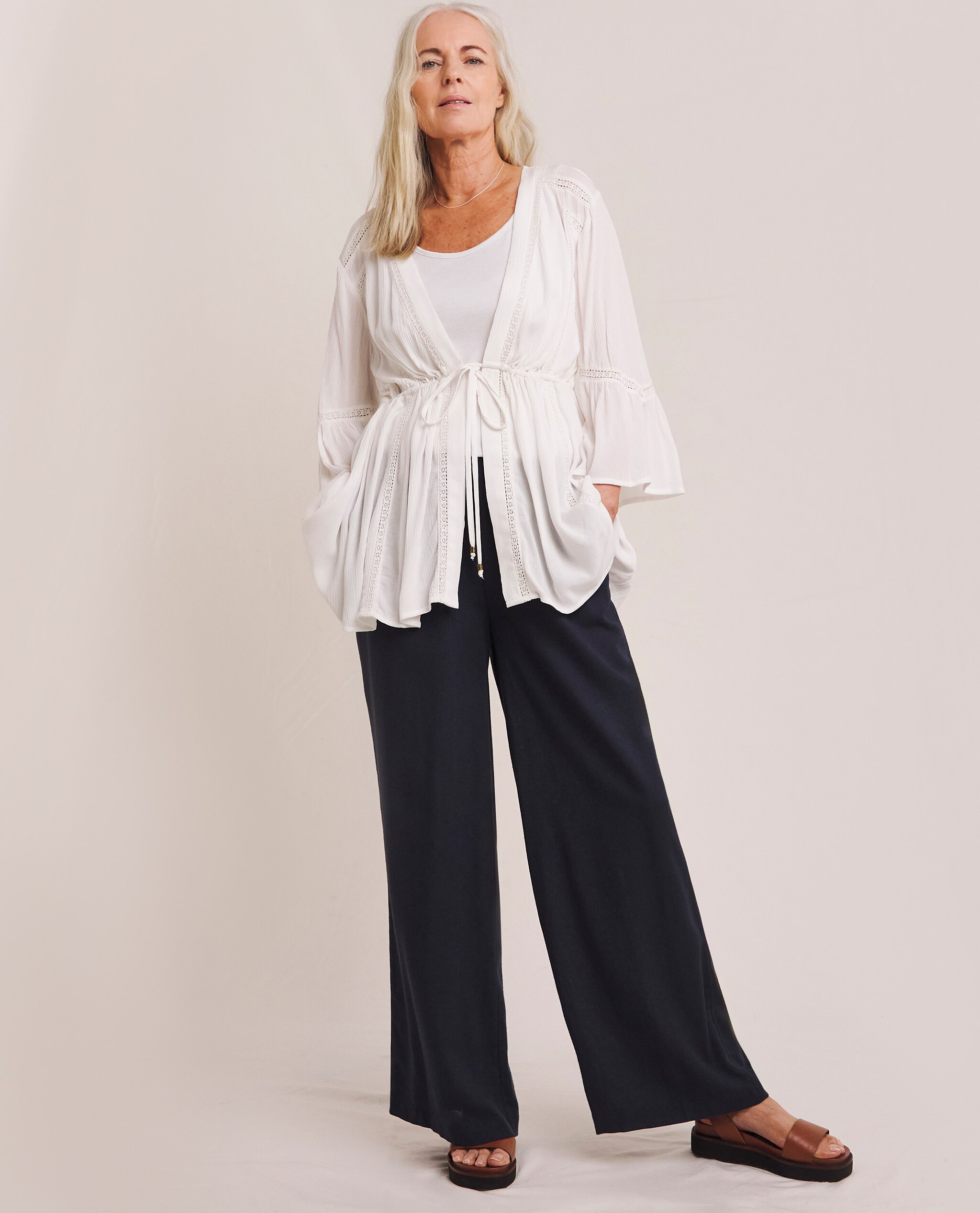 Model is wearing a crinkle lace trim tie front kimono with a pair of black trousers with tan sandals.