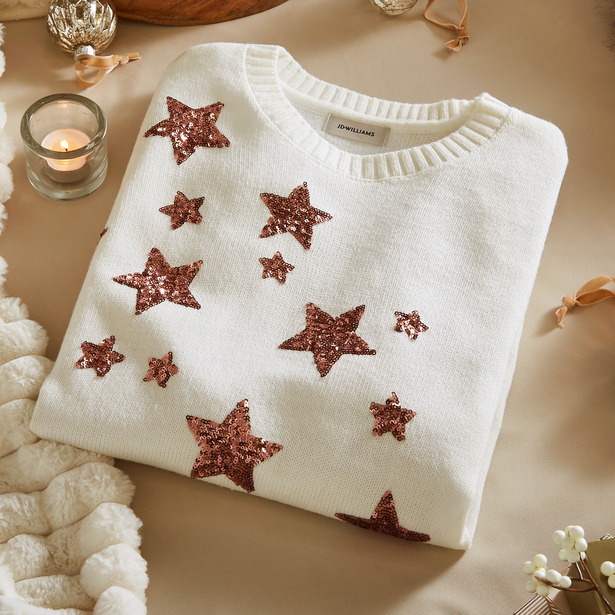 White jumper with glittery brown stars on a beige surface, surrounded by a candle and ornaments.