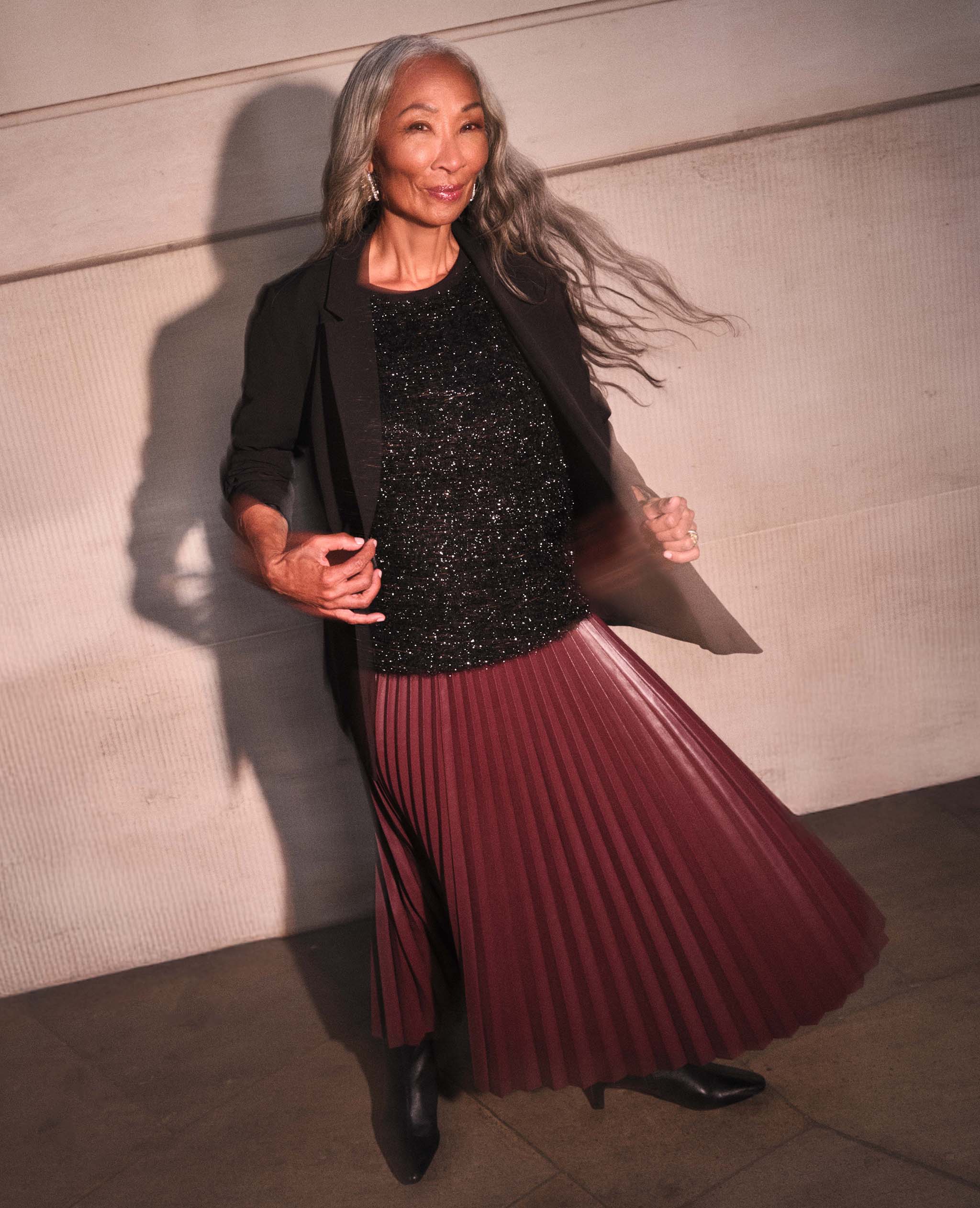 A lady outside wearing a black blazer a black sparkly top and a burgundy pleated skirt.