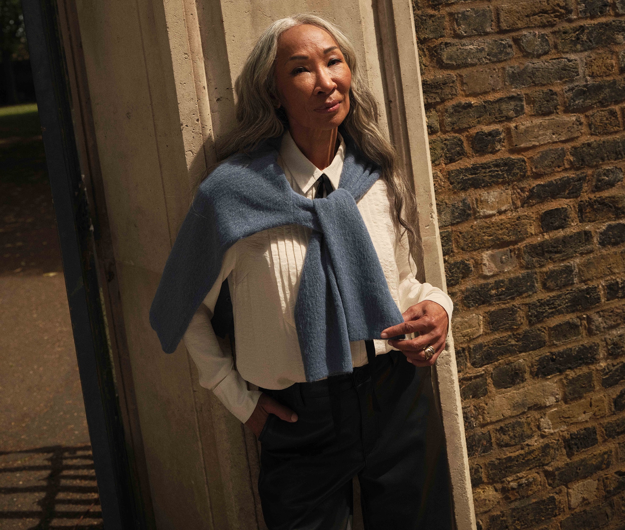 Lady walking down a street next to a brick wearing a white tuxedo shirt with black thin tie with a pale blue jumper draped over her shoulders
