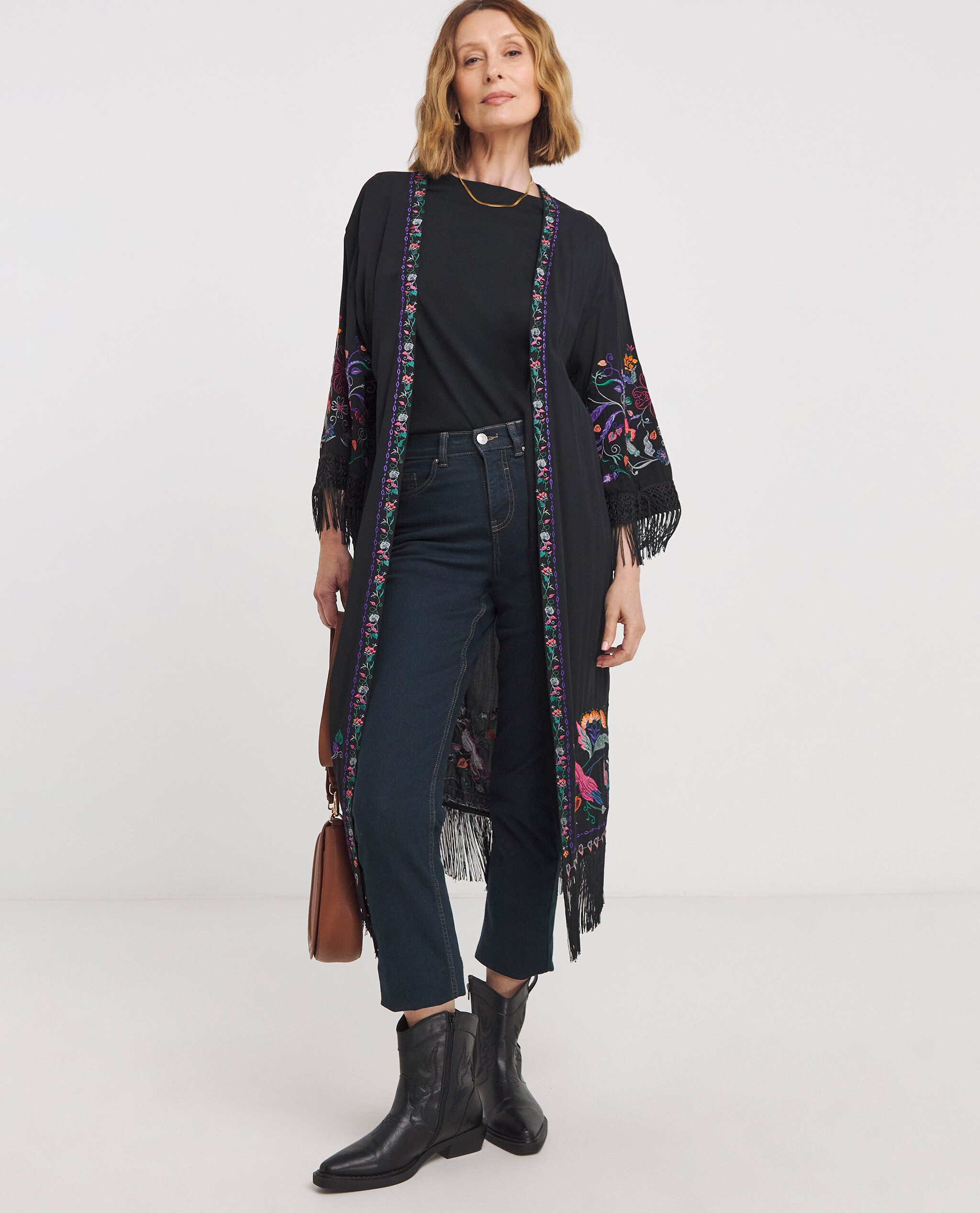 Model is wearing a black top underneath an embroidered kimono with dark denim jeans and black ankle boots with a tan handbag.