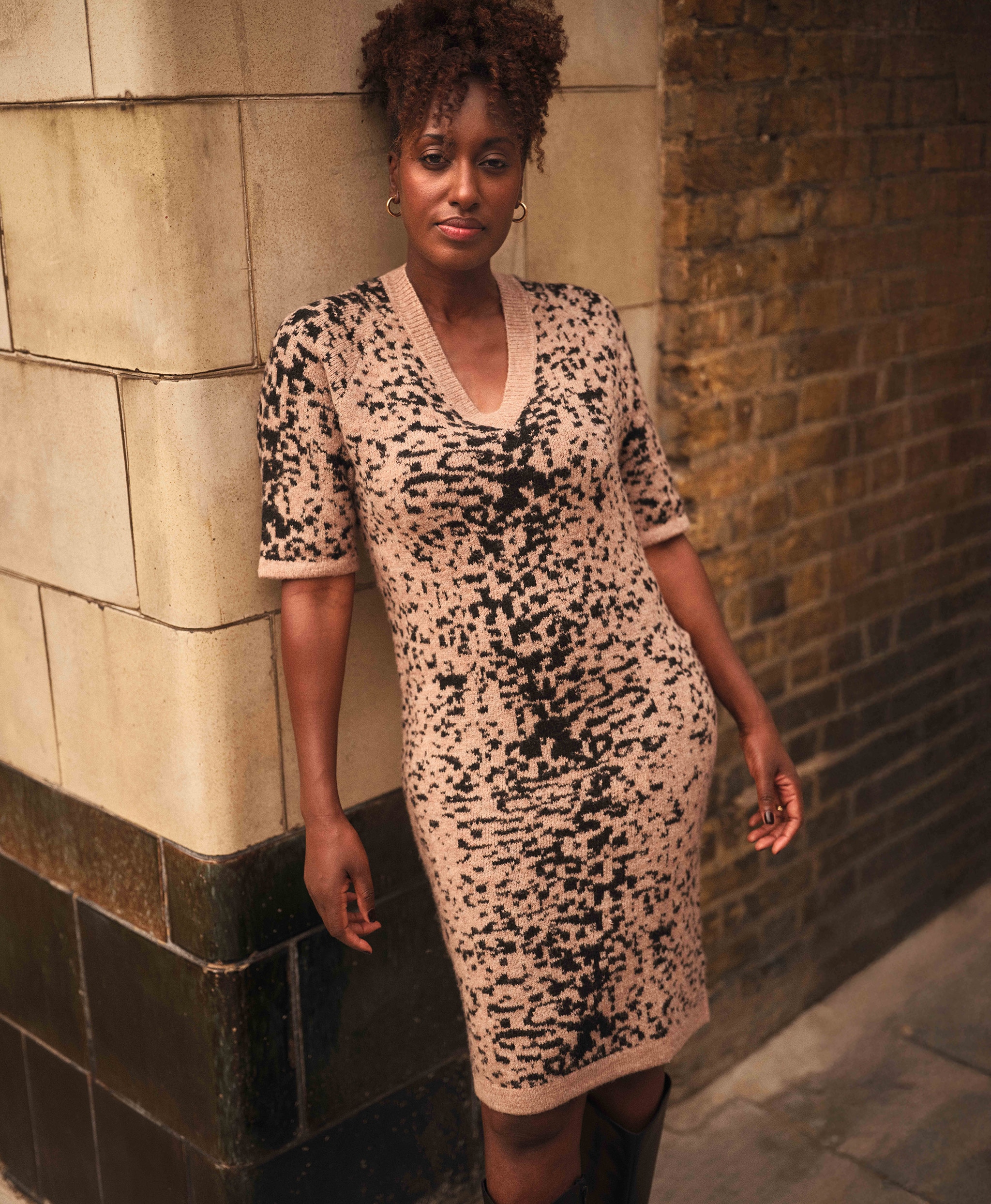 A lady stood on a street corner leaning against a wall wearing a knitted dress.