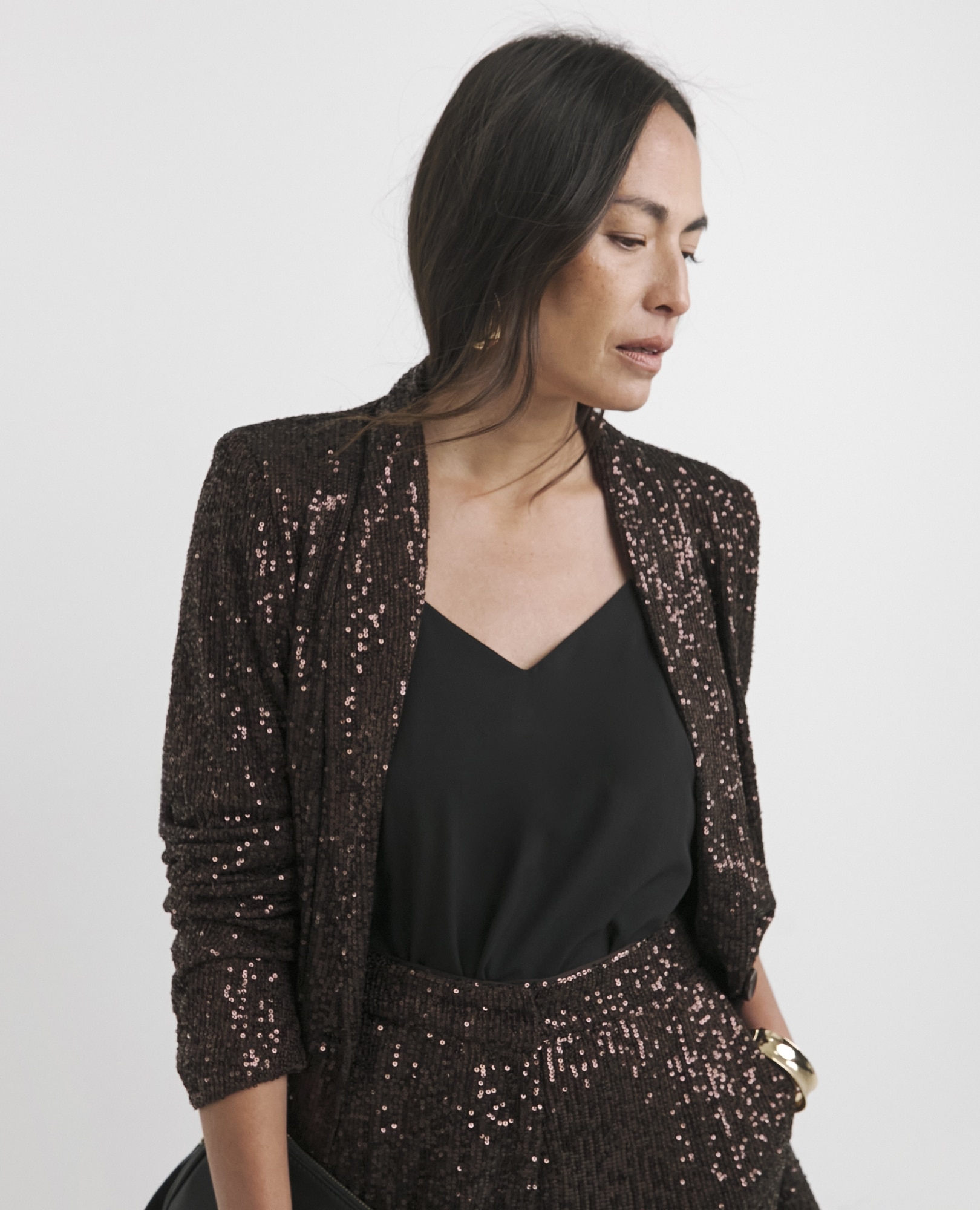 A woman with long dark hair wearing a sparkling brown sequin blazer and a black V-neck top, looking down