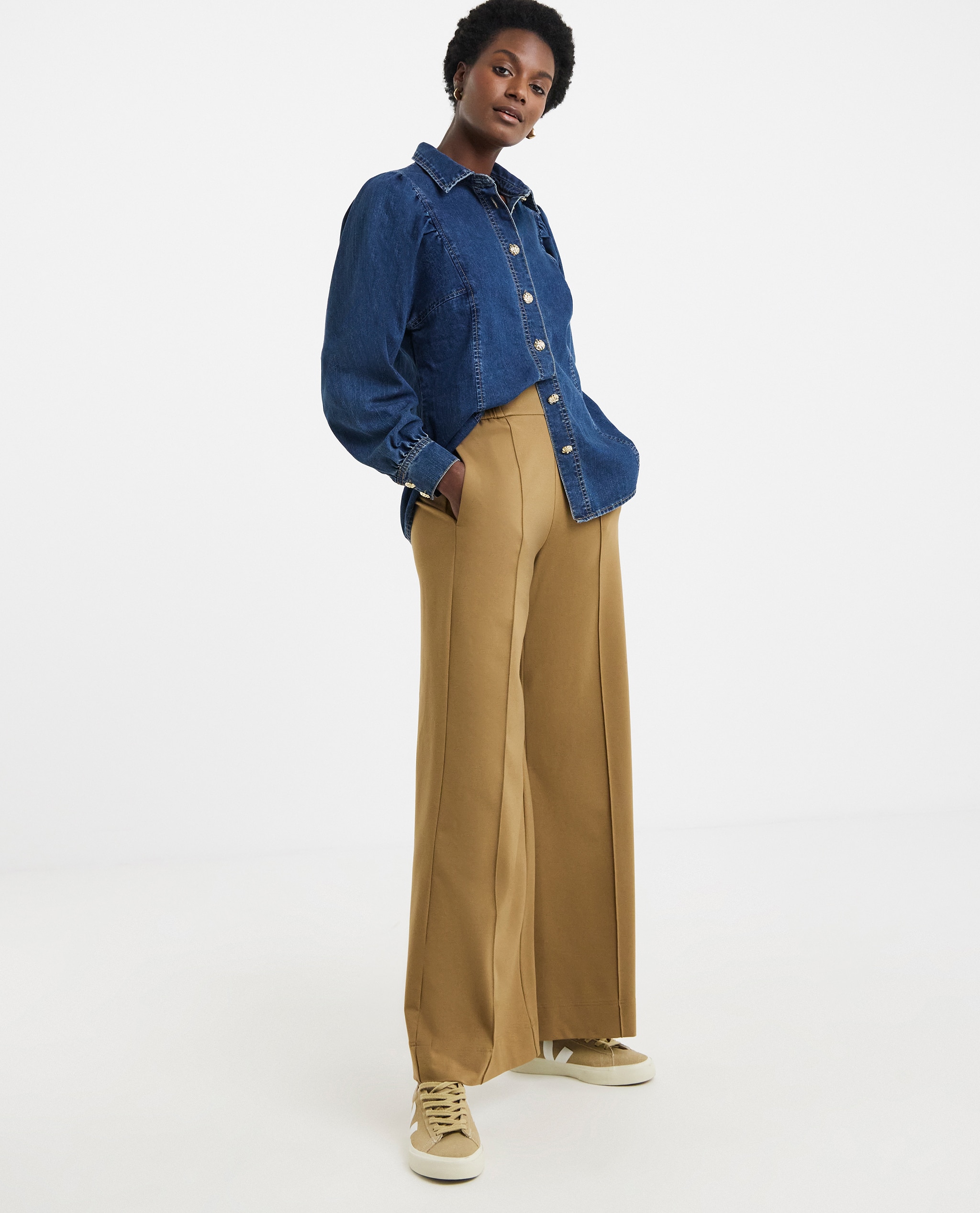 Model is wearing a mid blue volume sleeve denim top with a pair of camel twill straight leg trousers and trainers.