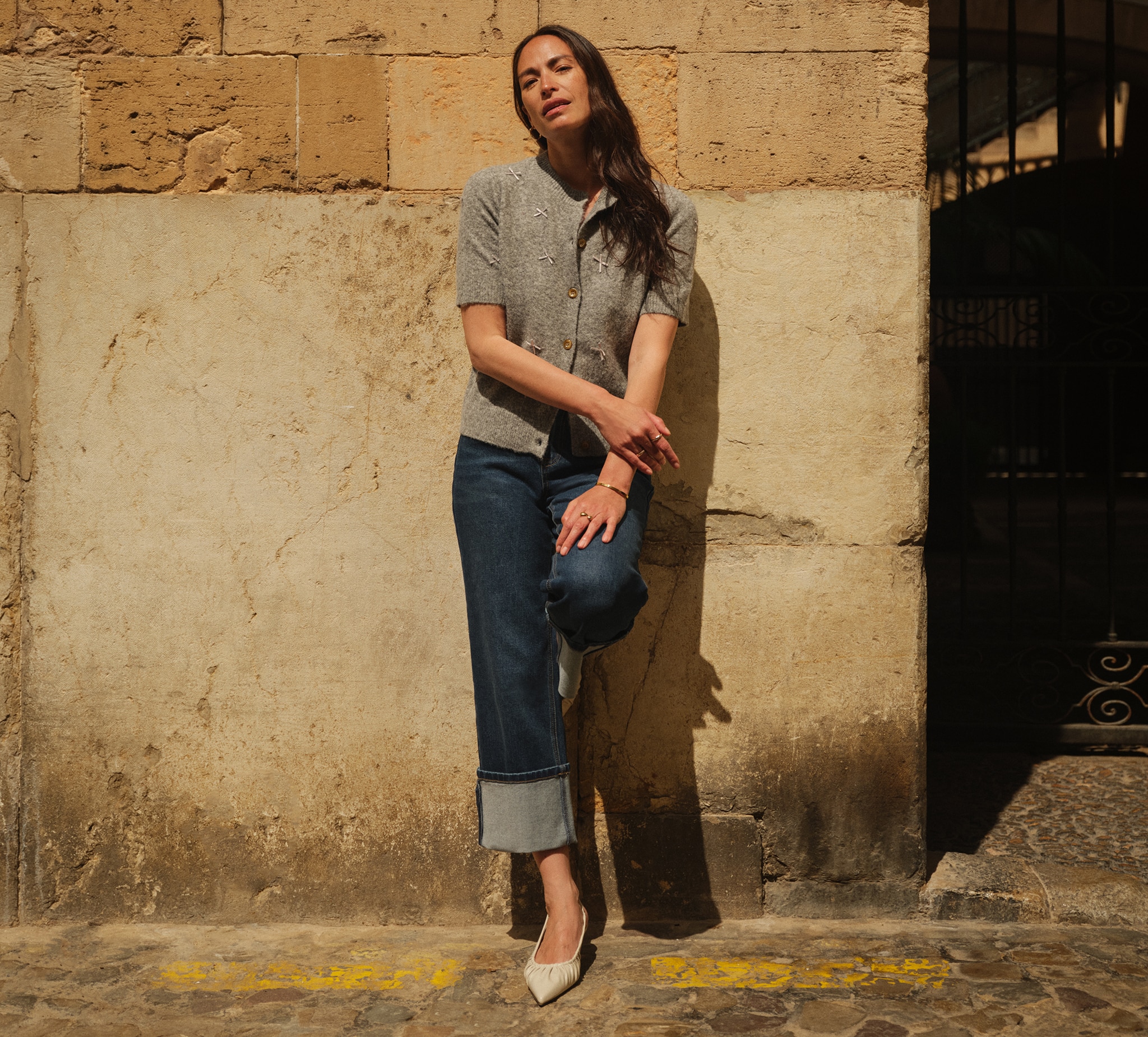A lady leaning on a sandy coloured stone all with one leg up wearing a grey short sleeved top and blue jeans.