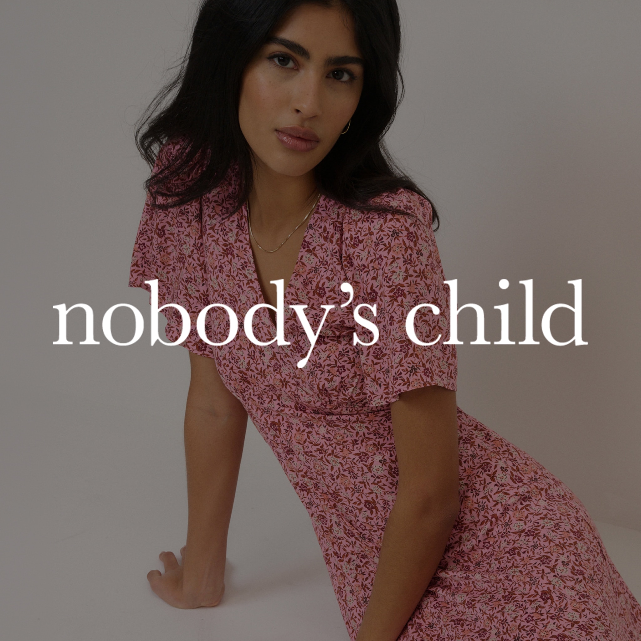 A lady sat on the floor wearing a pink Nobody's Child dress. The Nobody's Child logo is overlaid on the image.