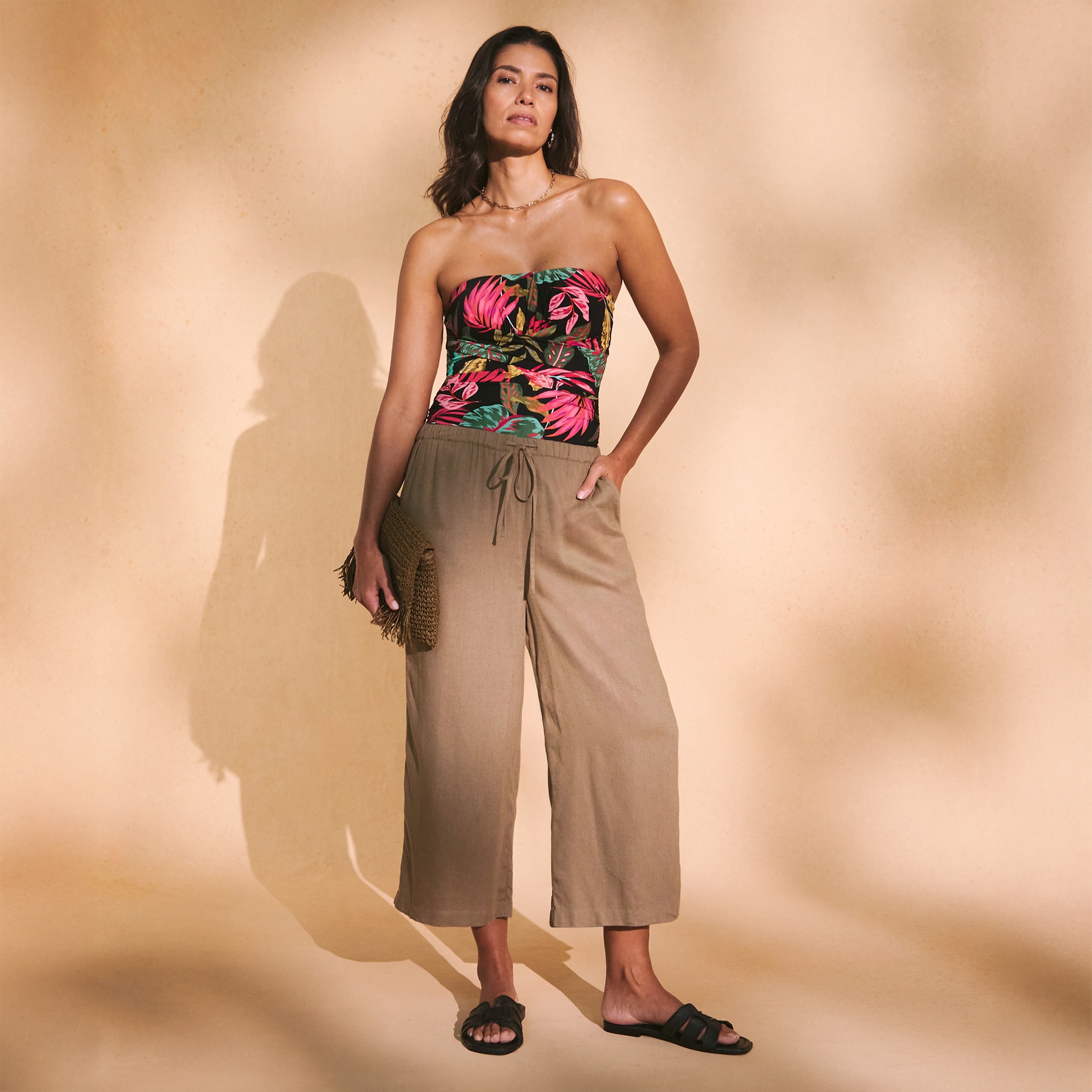A person is standing against a beige background wearing a strapless top with a vibrant tropical floral pattern, paired with linen trousers.