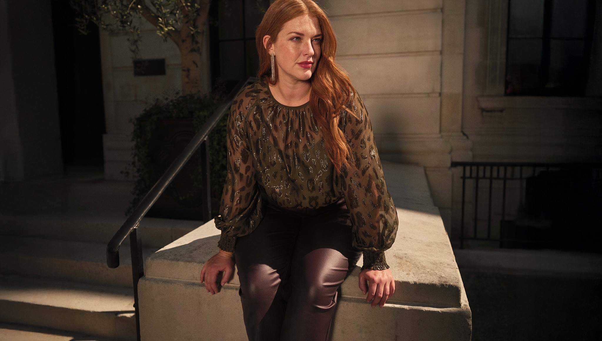 A lady sat on a wall outside wearing a green sparkly top with brown trousers