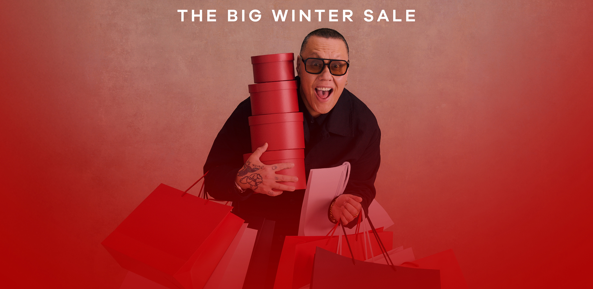 Gok Wan holding lots of shopping bags and parcels on a red background.
