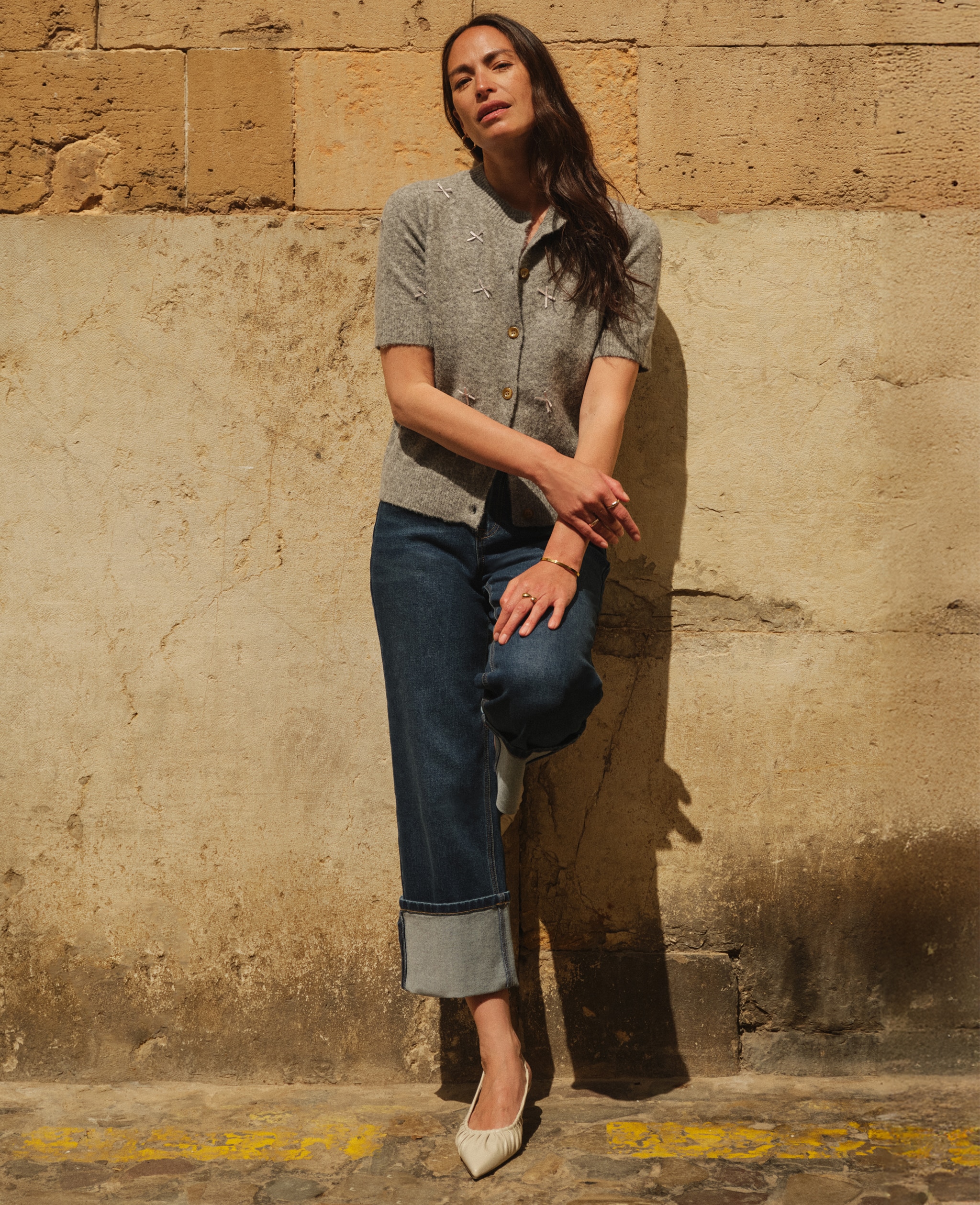 Lady wearing jeans and a grey top leaning against a wall.