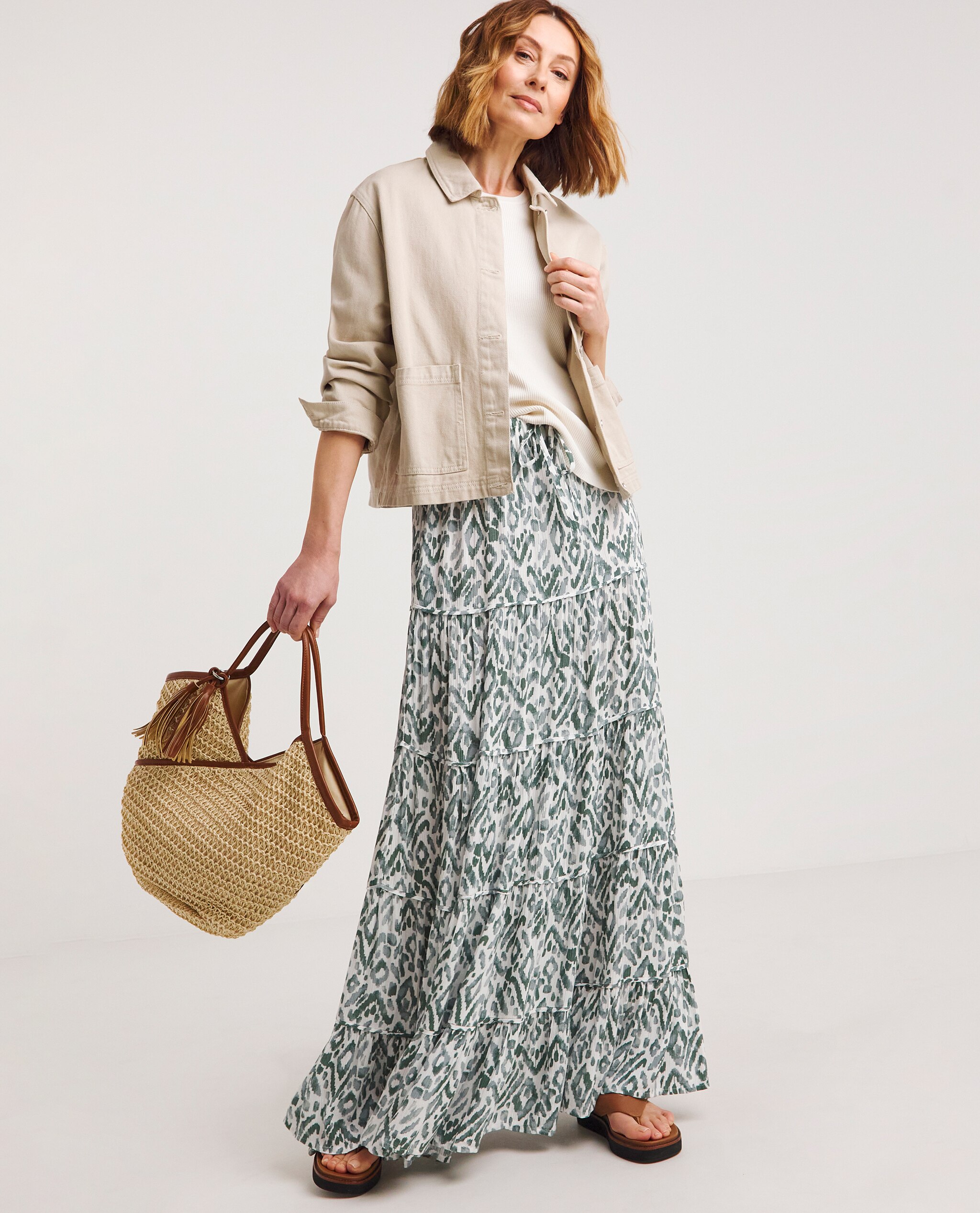 Model is wearing a cream denim jacket with a cream jumper underneath with a green print maxi skirt and tan toe post sandals and tote bag.