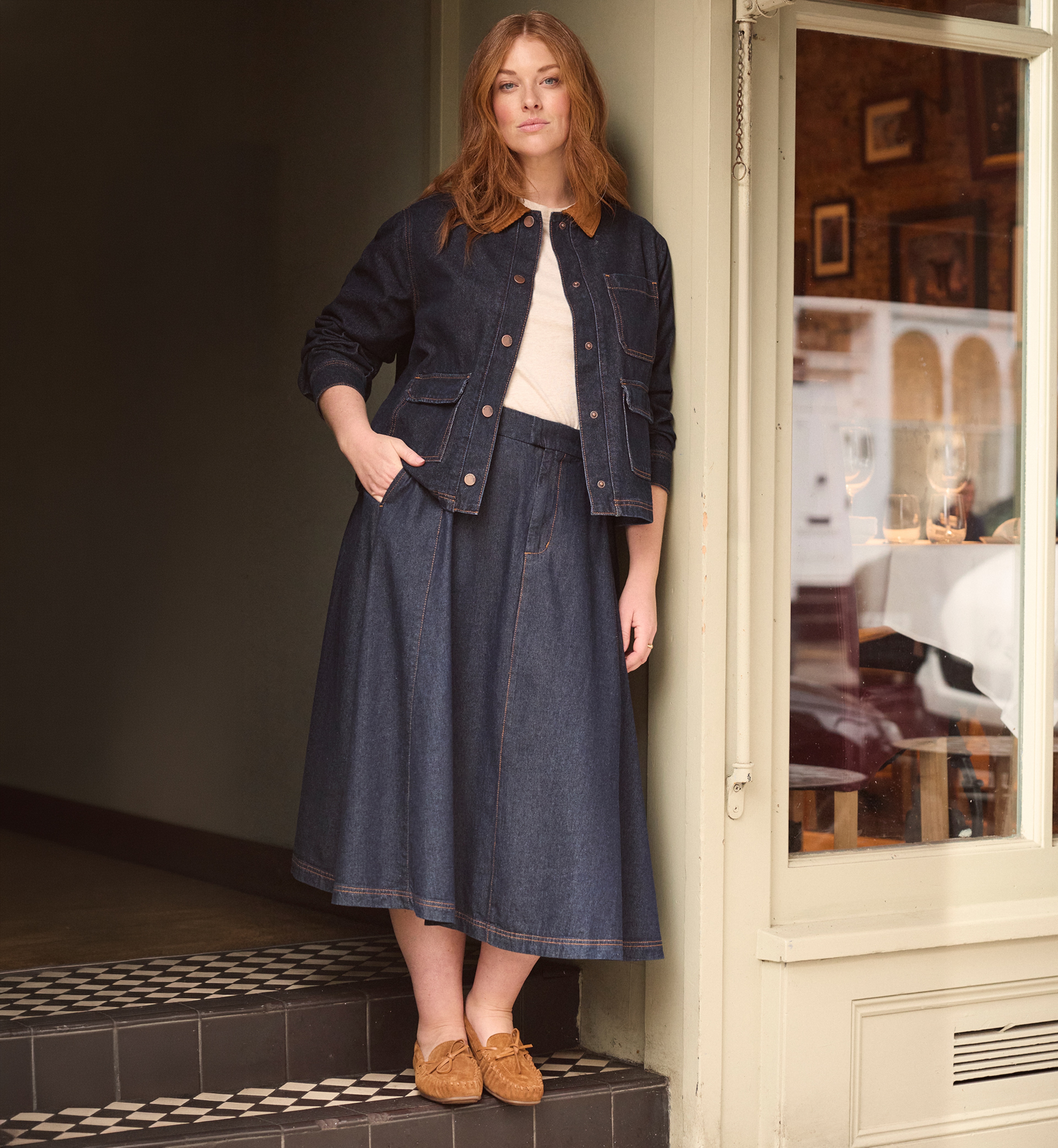 A lady stood in a door way wearing a denim jacket and denim skirt.