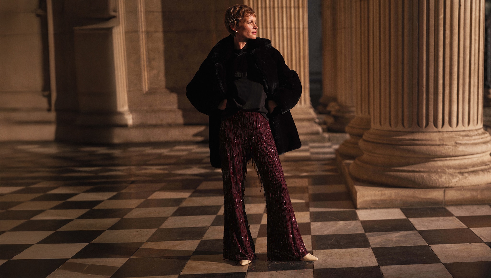 A lady stood outside in a big entrance next to big pillars and a chequered floor wearing glittery burgundy trousers a black top and a faux fur coat.