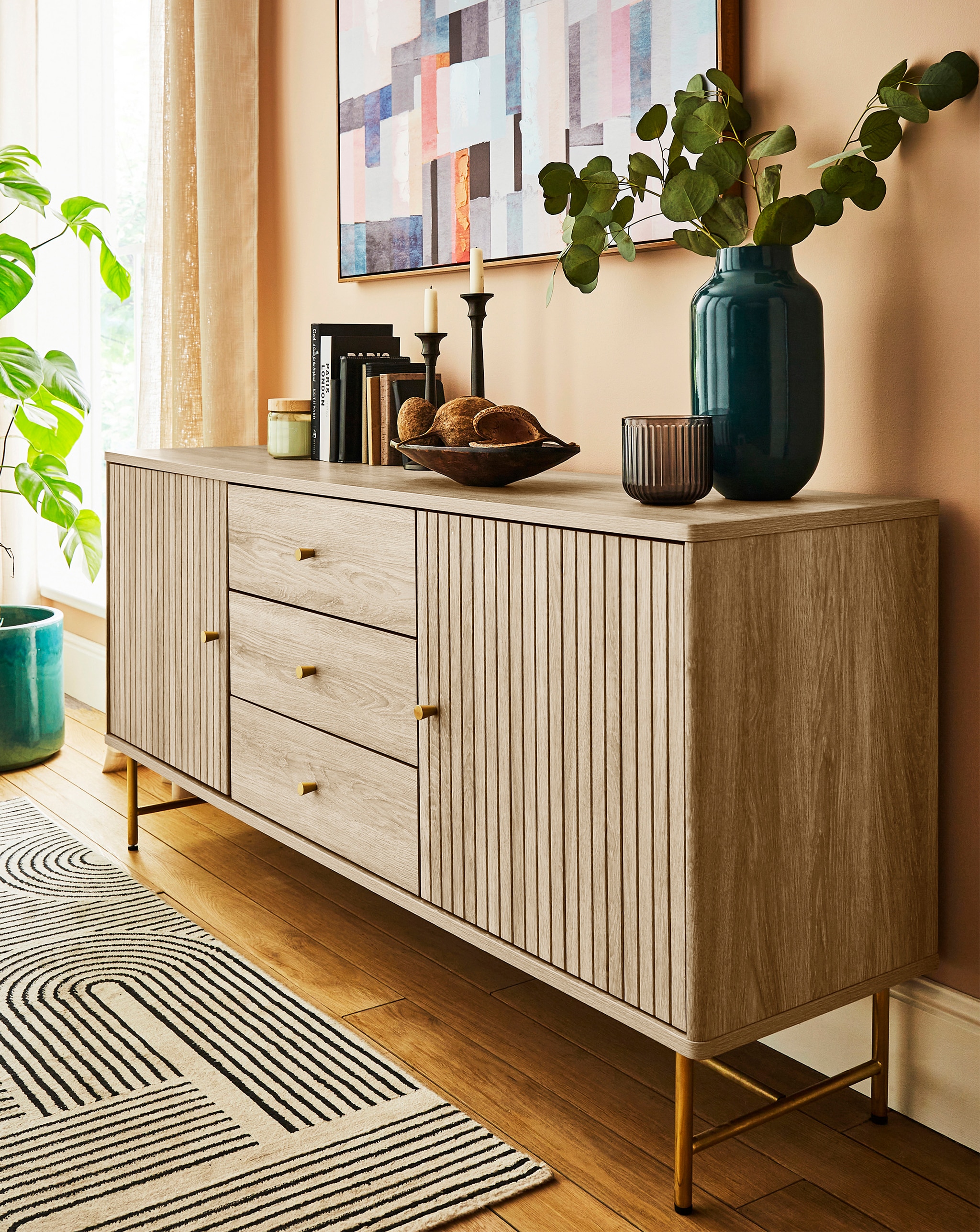 A modern living room featuring a light wood sideboard decorated with candles, books, a bowl, and a tall vase.