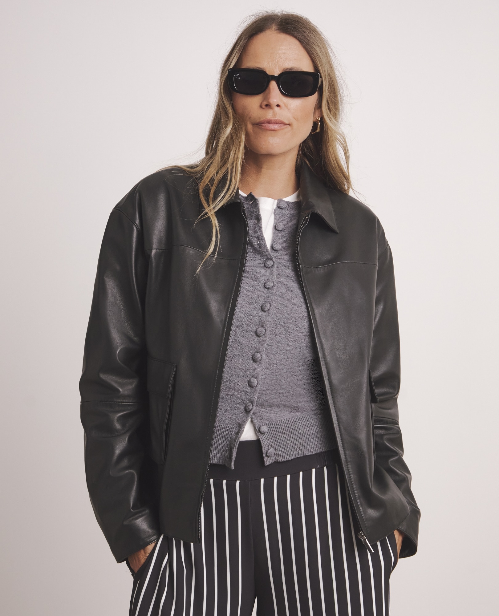 A stylish woman wearing black sunglasses, a dark leather bomber jacket, a grey cardigan, and black and white vertically striped trousers