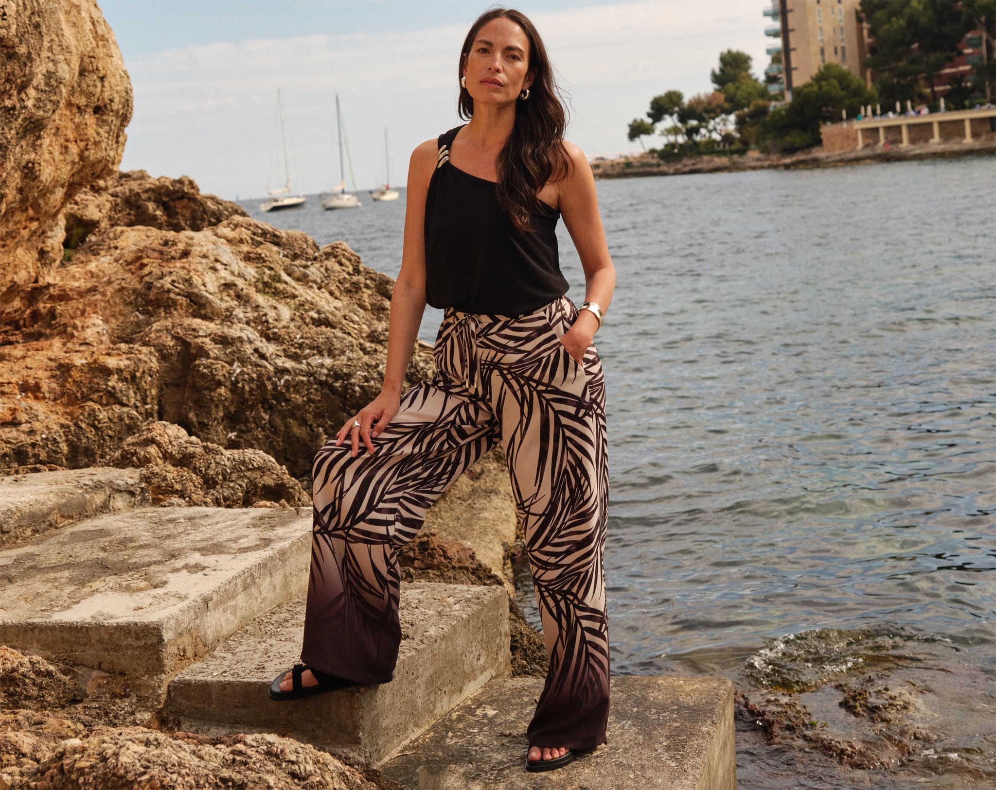 A lady wearing a black vest top and gold and black animal print trousers stood by the sea.