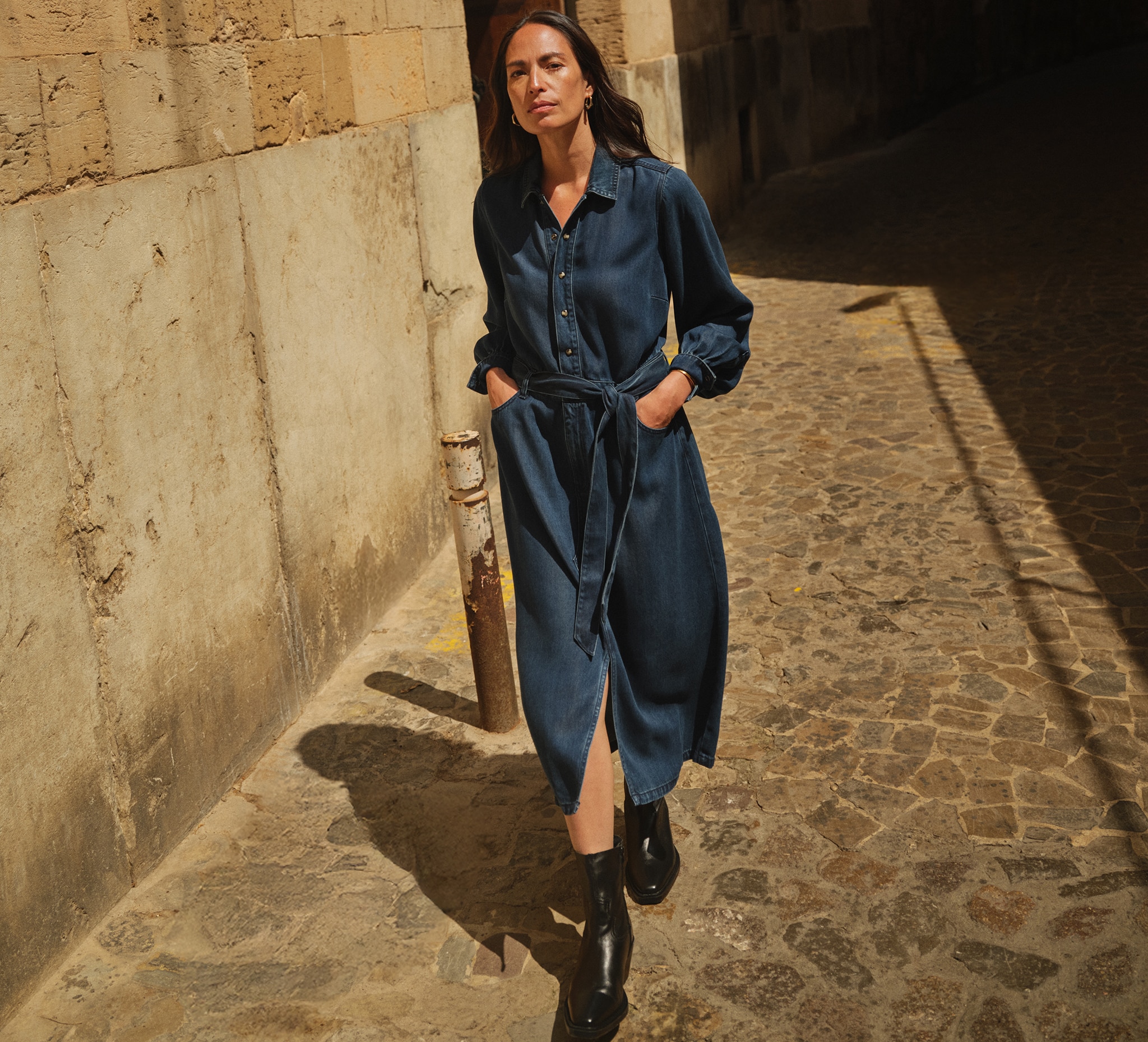 A lady wearing a denim dress walking on a cobbled street with stone walls.