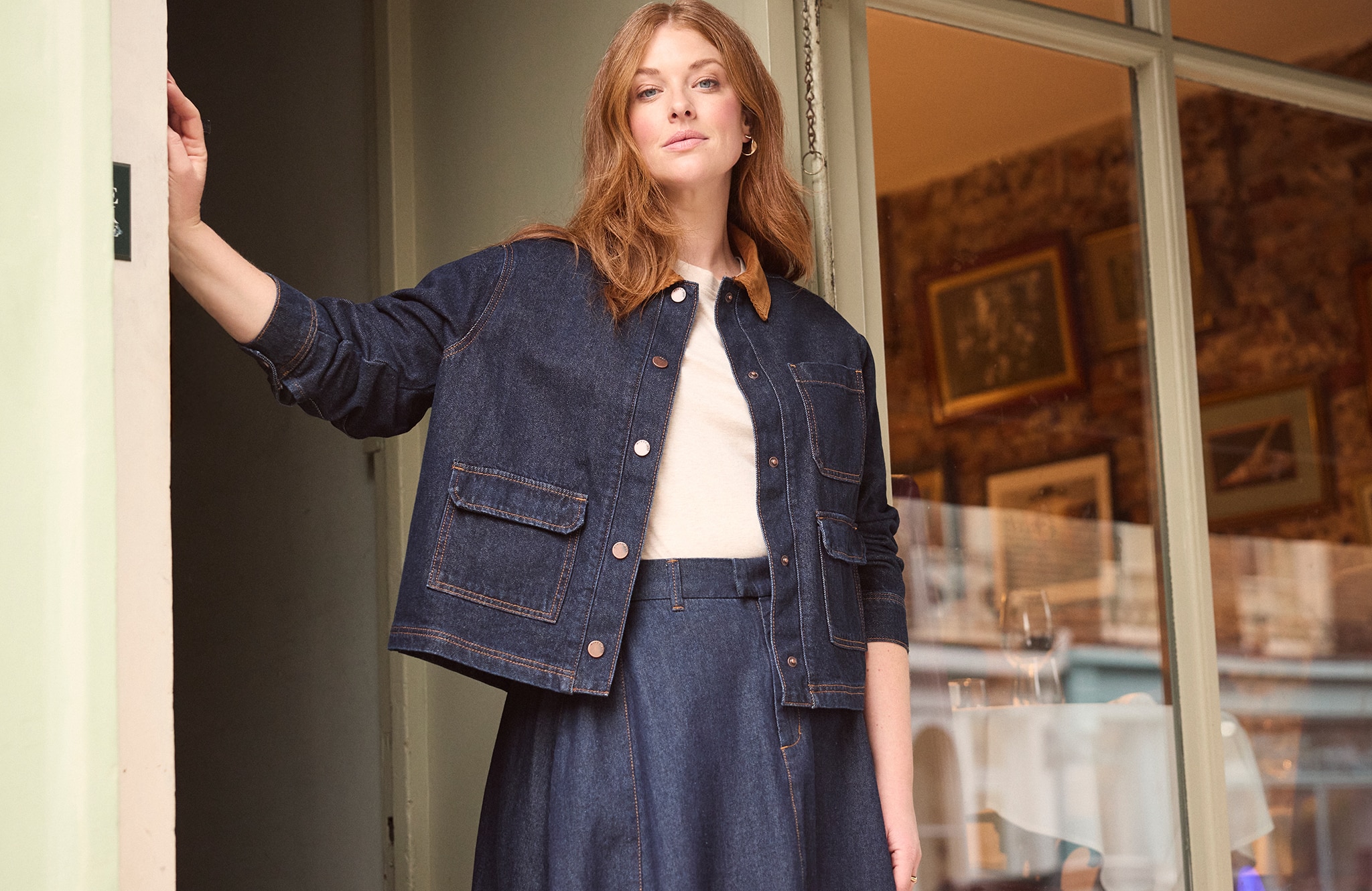 Model is wearing a indigo cord collar denim barn jacket over a cream top with an indigo seamed full circle midi skirt.