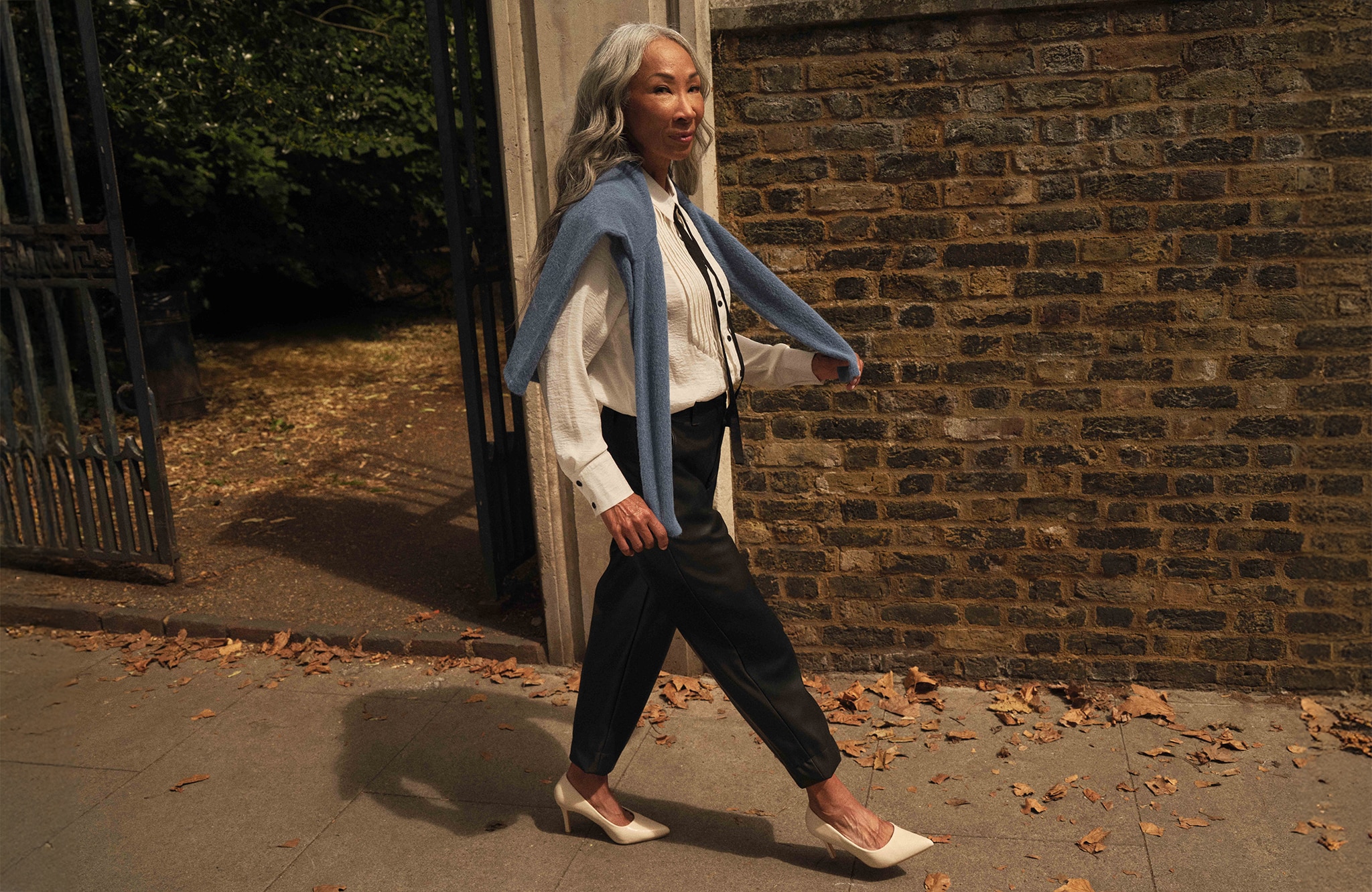 Lady walking down a street next to a brick wearing a white tuxedo shirt with black thin tie with a pale blue jumper draped over her shoulders