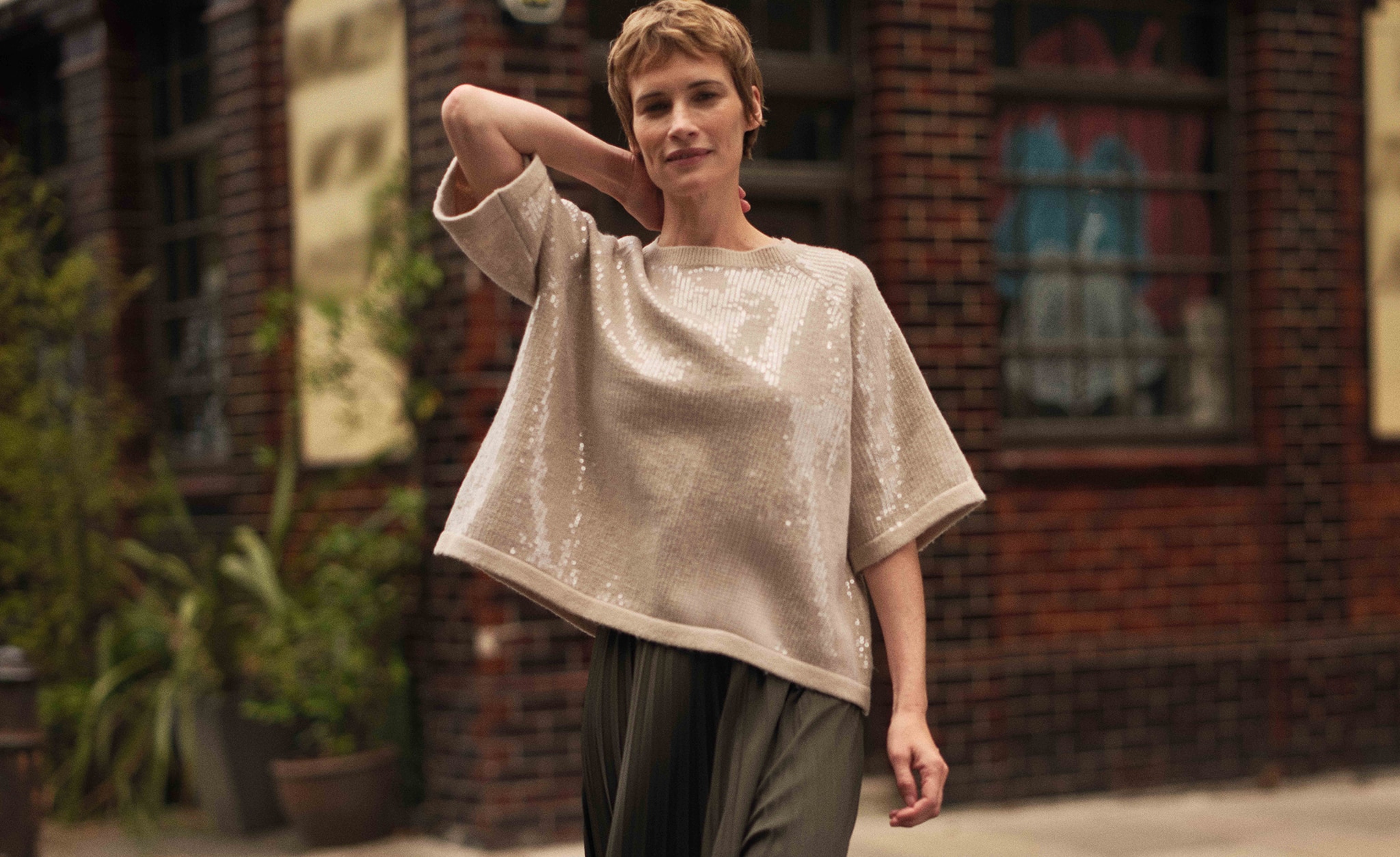 A lady walking outside stretching her arm round the back of her head wearing a gold / champagne coloured sequin top.