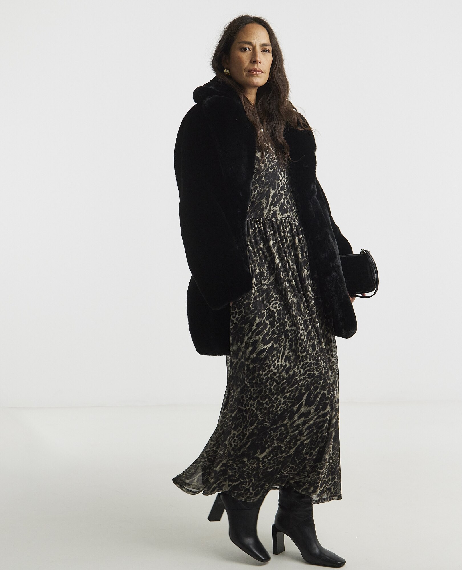 A woman with long dark hair wearing a black faux fur coat over a long leopard-print maxi dress, paired with black mid-calf boots