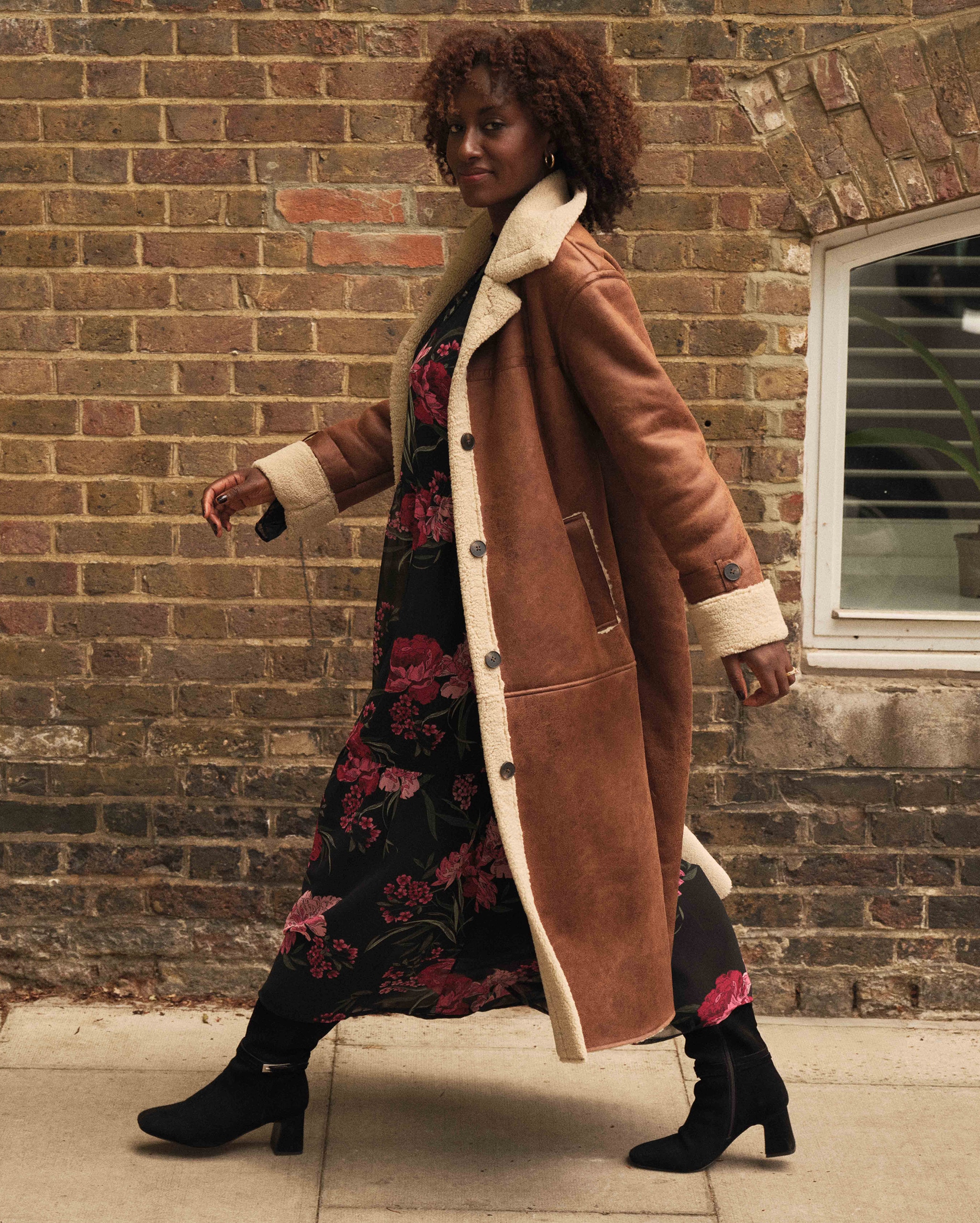 A woman walking right to left on a street wearing a tan maxi shearling coat.