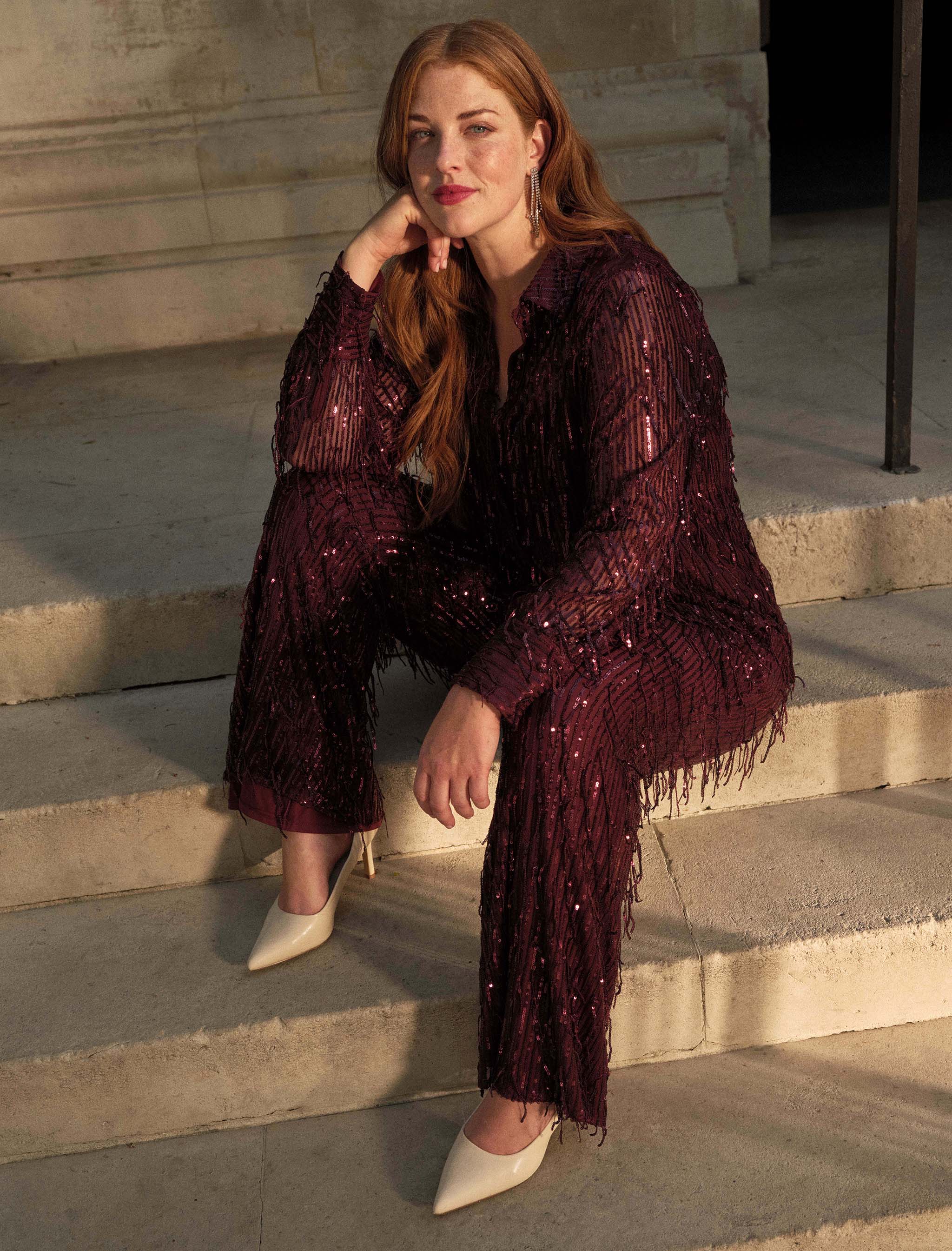 A lady sat on some steps wearing matching burgundy sequin top and trousers.