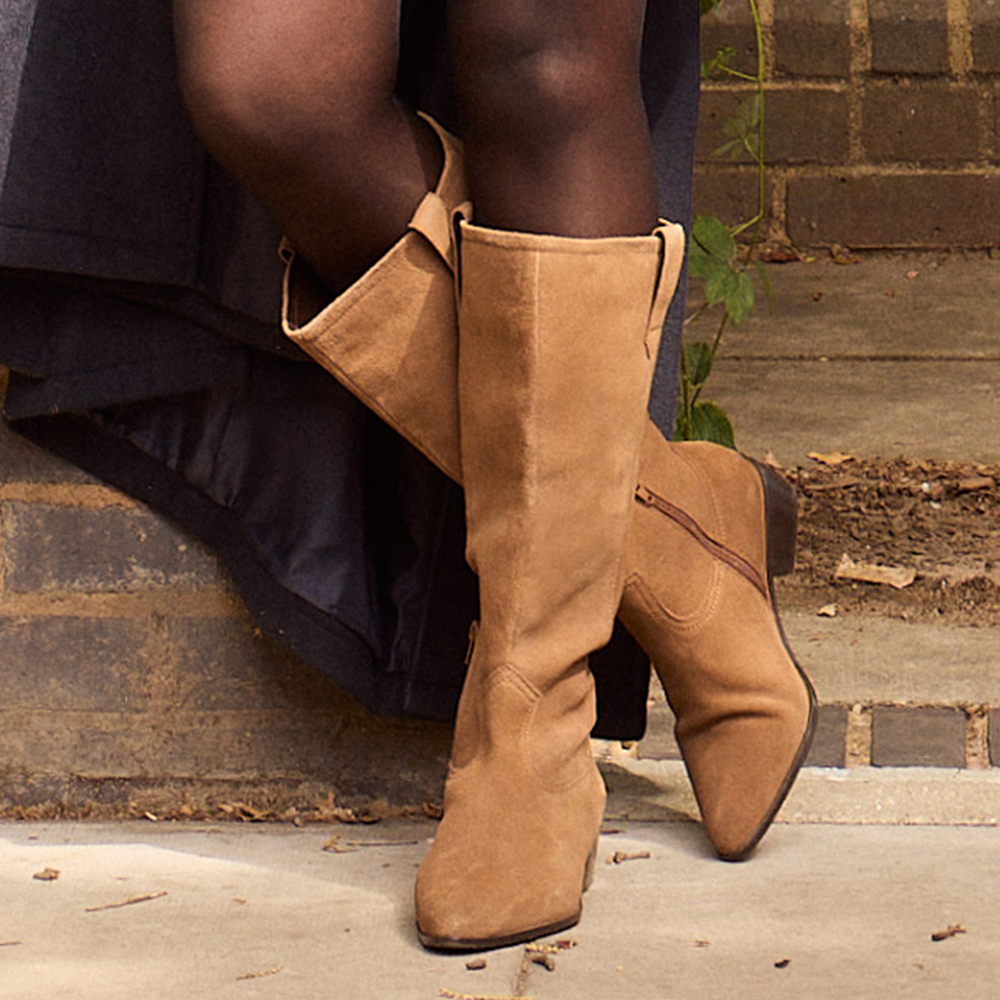A person wearing browny tan coloured knee high boots stood still on a pavement.