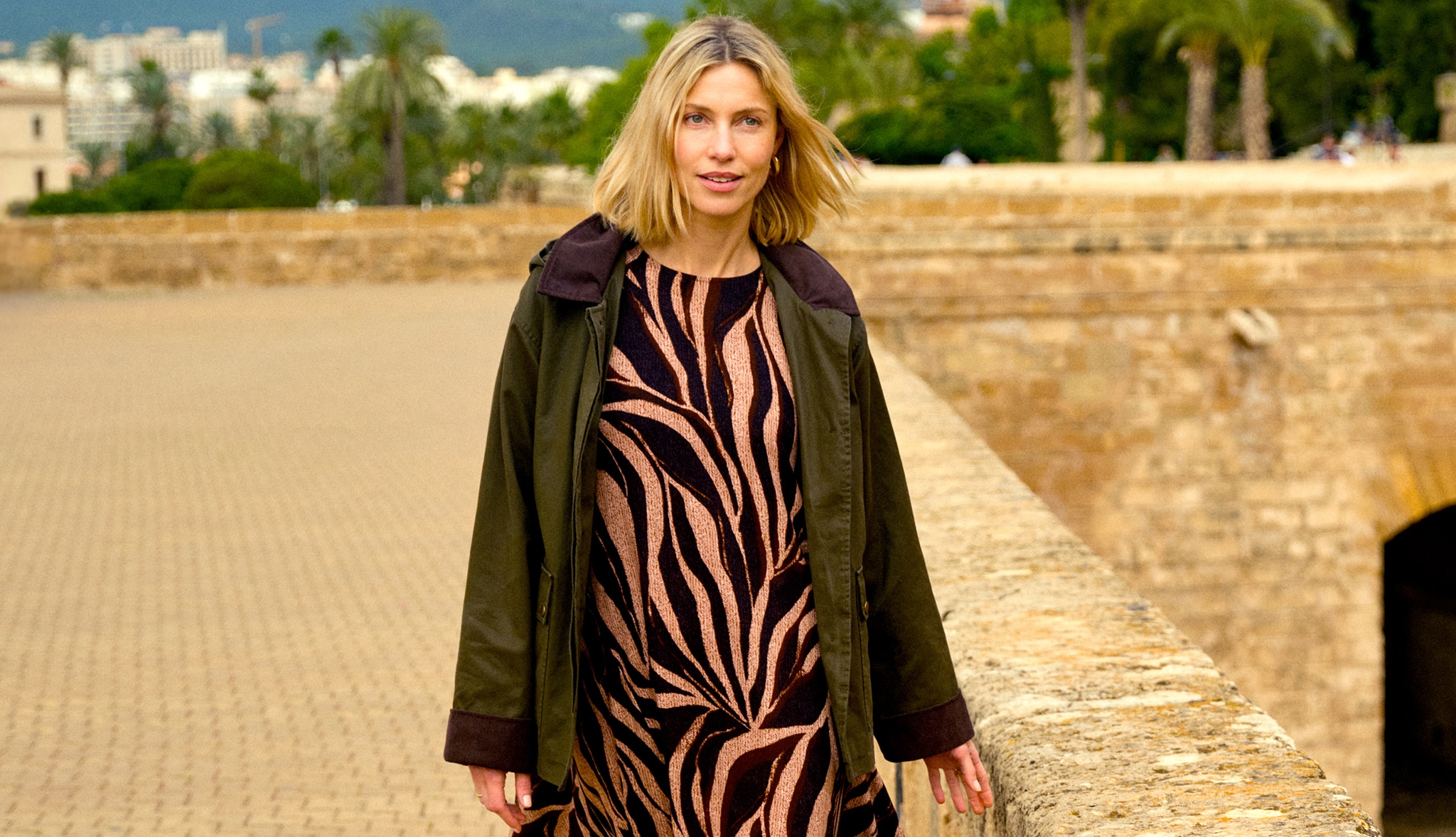 A lady walking next to a stone wall wearing a bronze an black animal print dress.