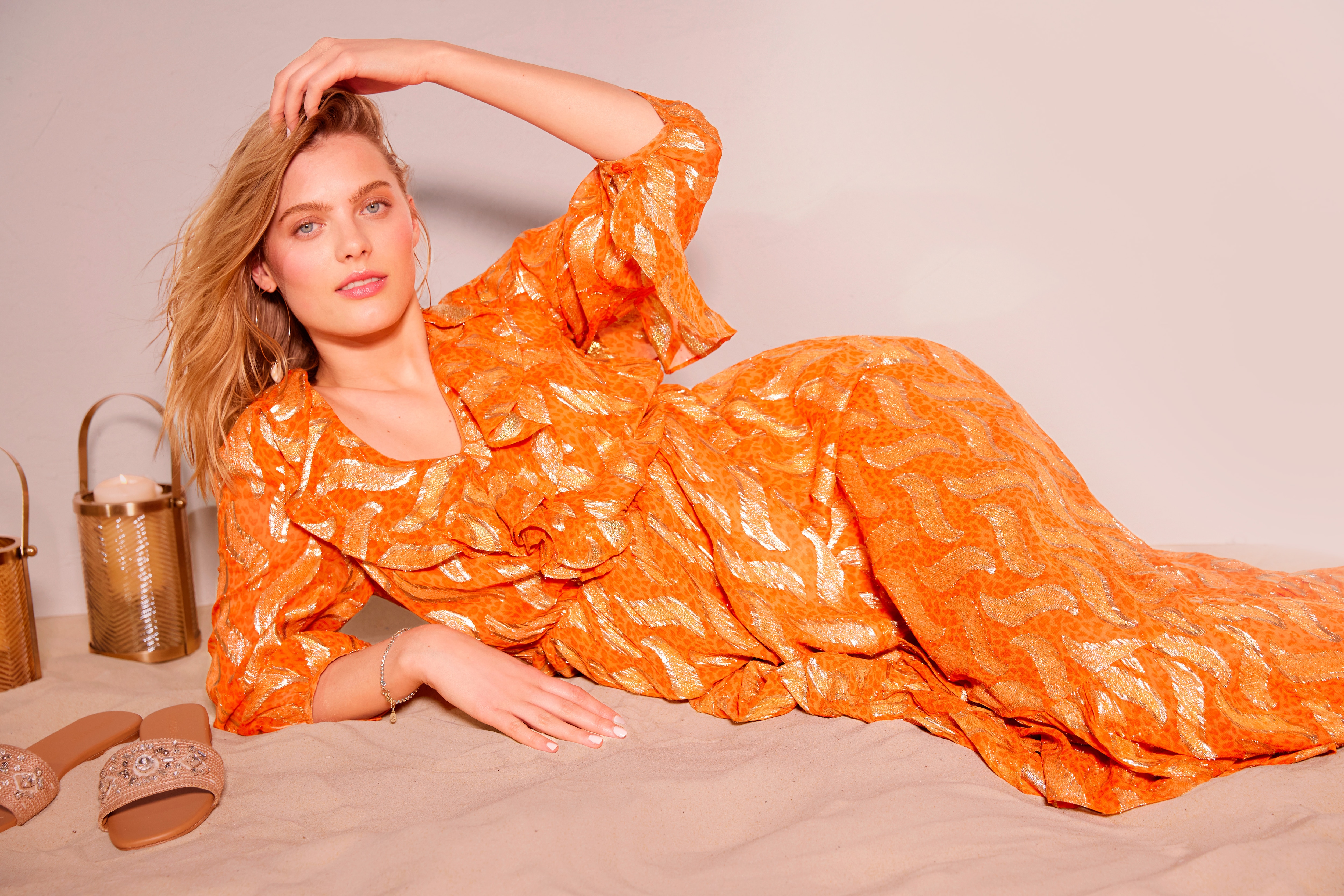 Model reclining on sand with sandals and wearing  an orange summer dress with gold detail.