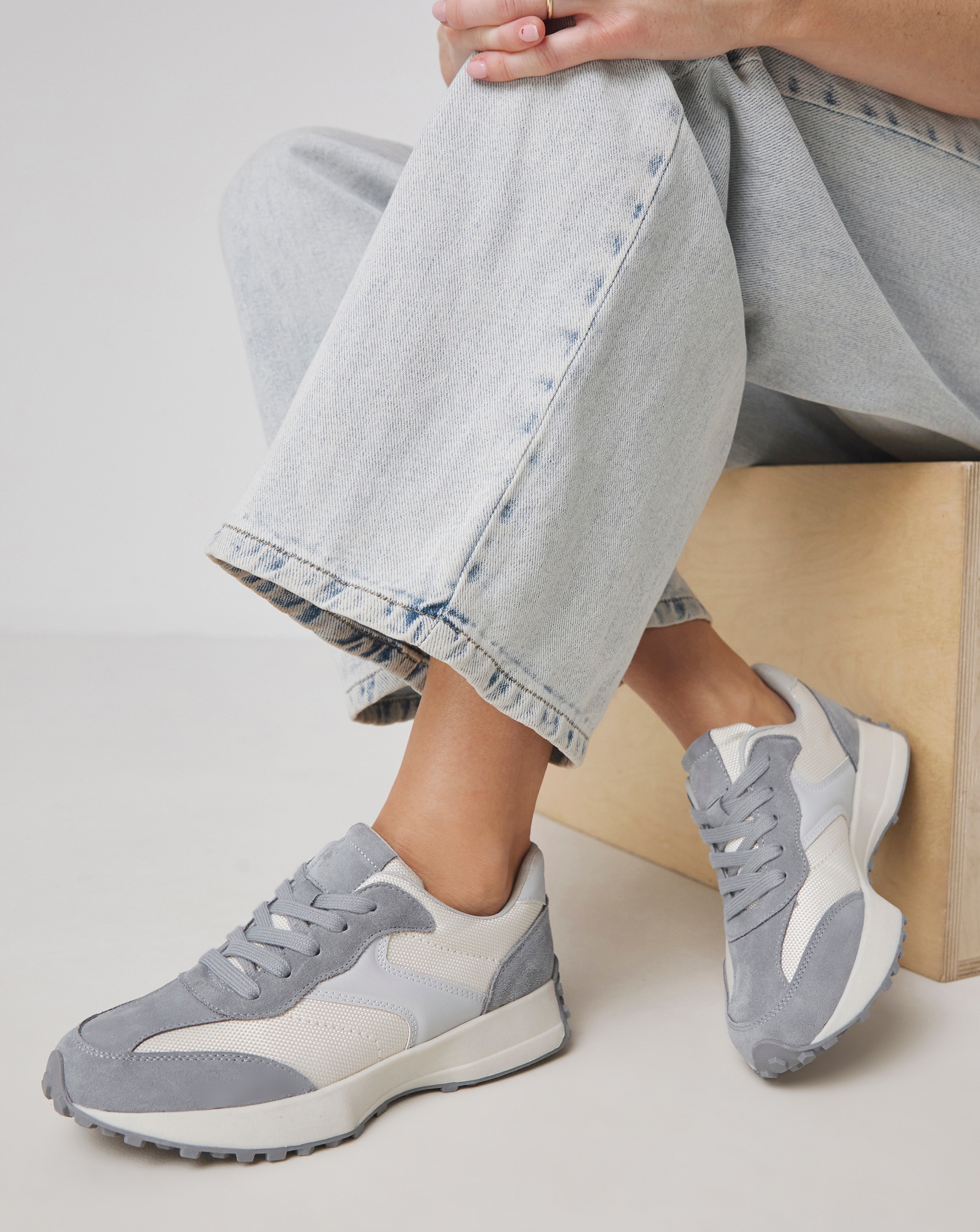 A person sat on a wooden box wearing pale blue denim jeans with Grey panelled trainers