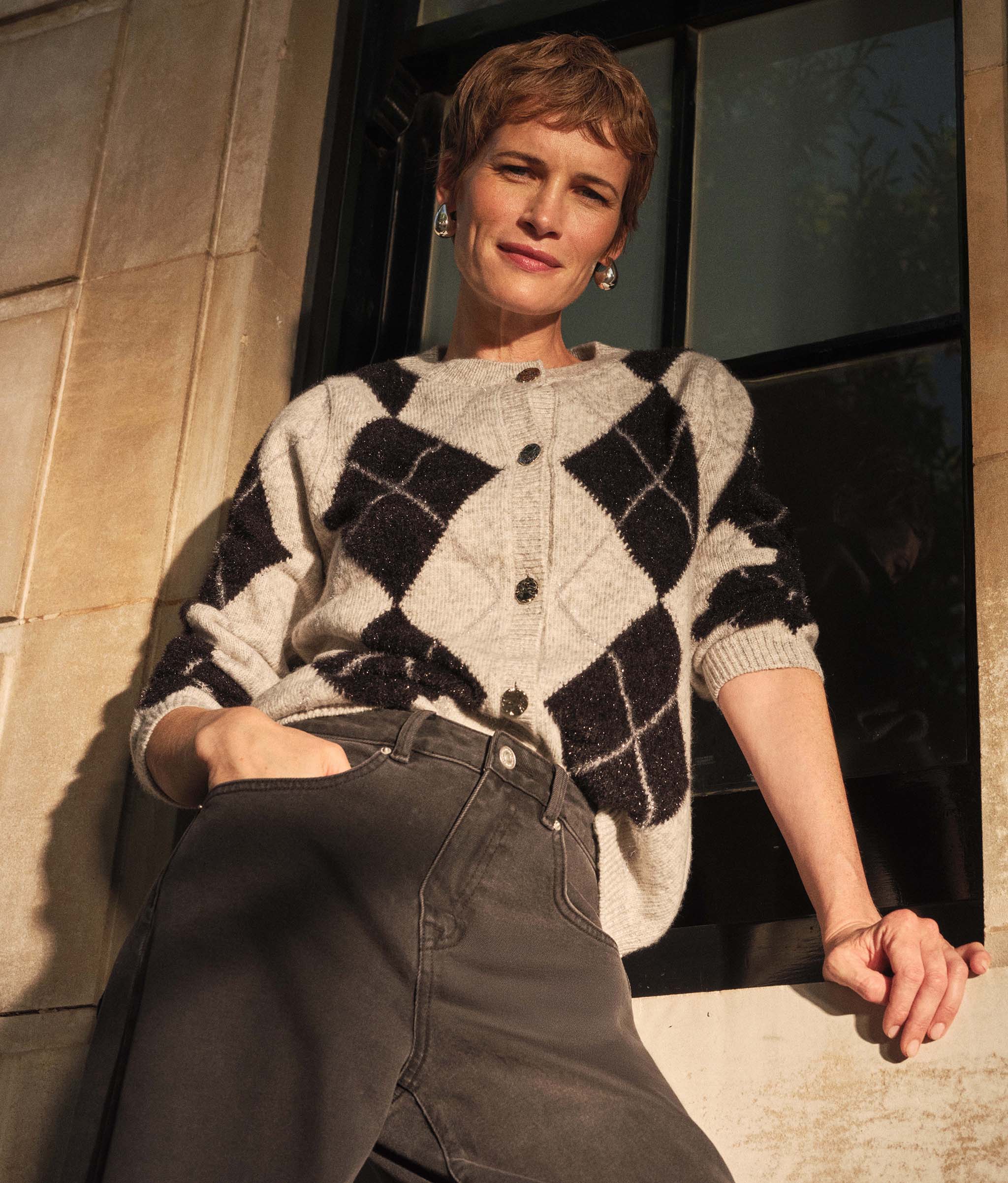 A lady stood outside leaning against a wall wearing a grey and black argyle cardigan with black jeans.