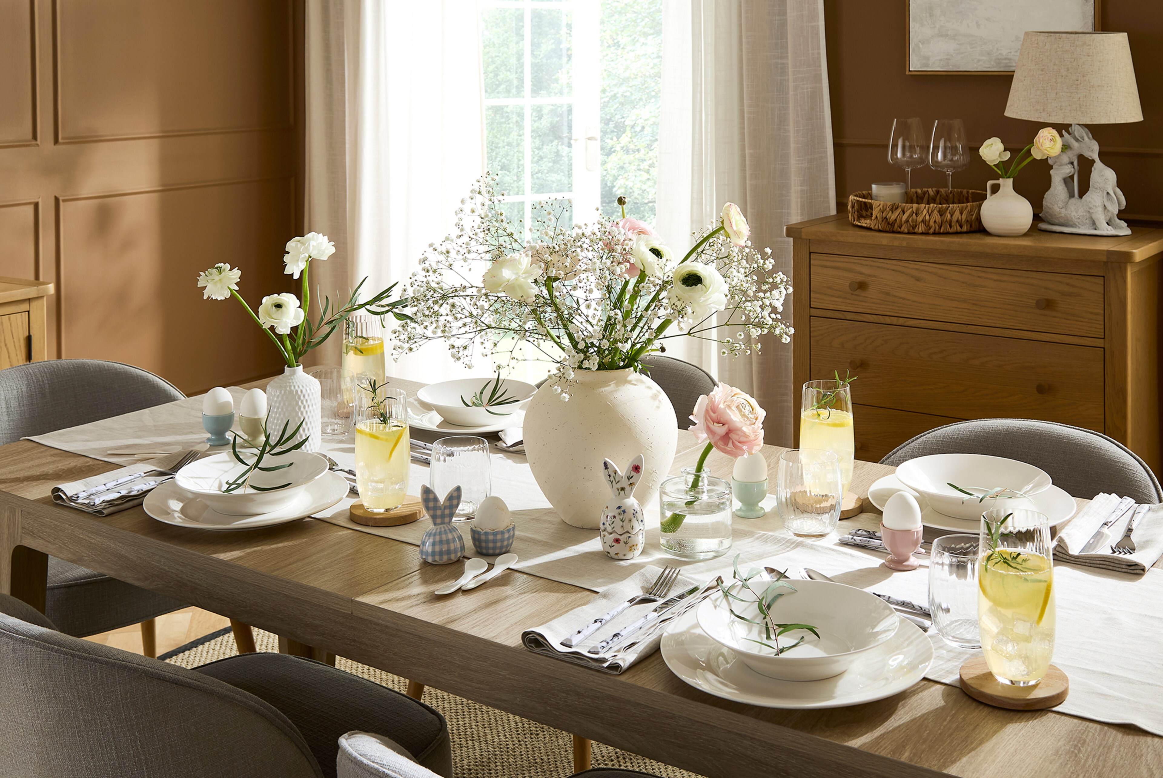 Julipa Charlotte extendable dining table set for a meal with white plates, glasses and small decorative bunny figurines in a bright, modern dining room.