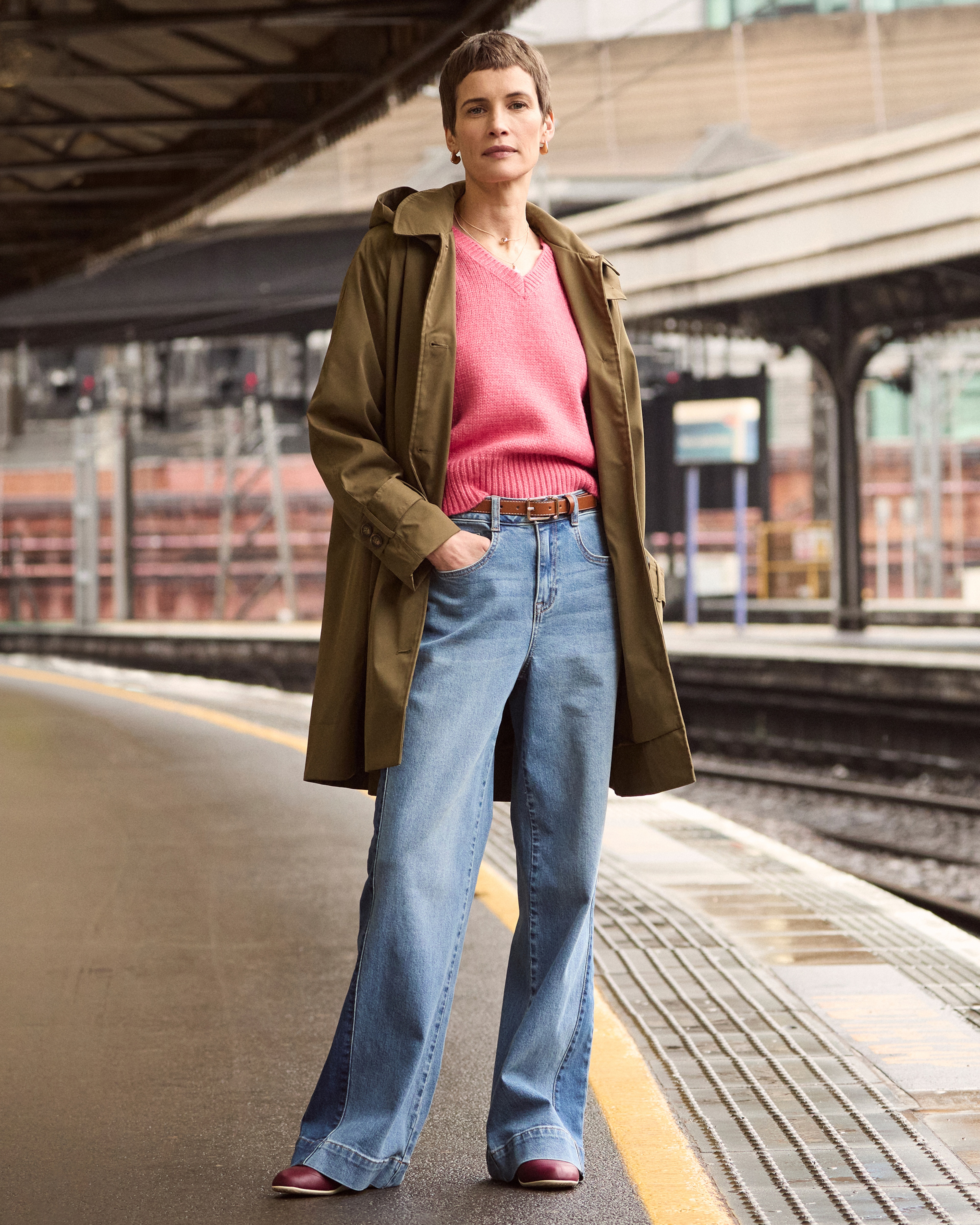 A lady stood in a train station wearing pale blue jeans, a pink jumper and Khaki coat.