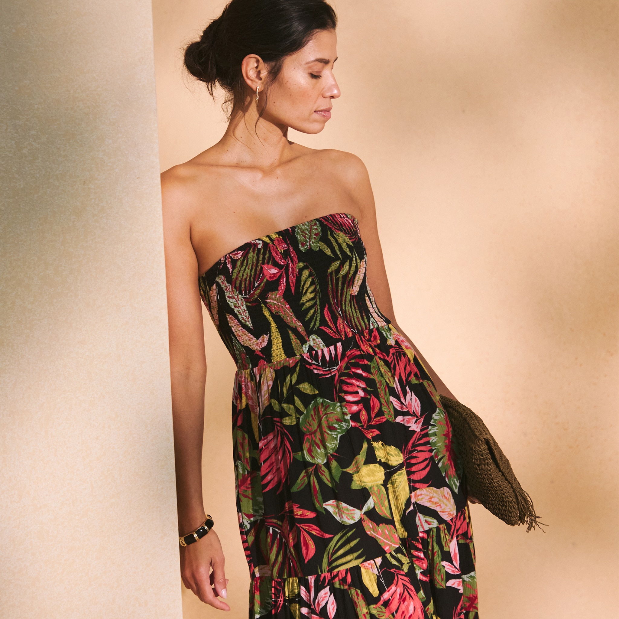A person wearing a strapless black maxi dress with a colourful tropical leaf print, holding a brown textured clutch bag.