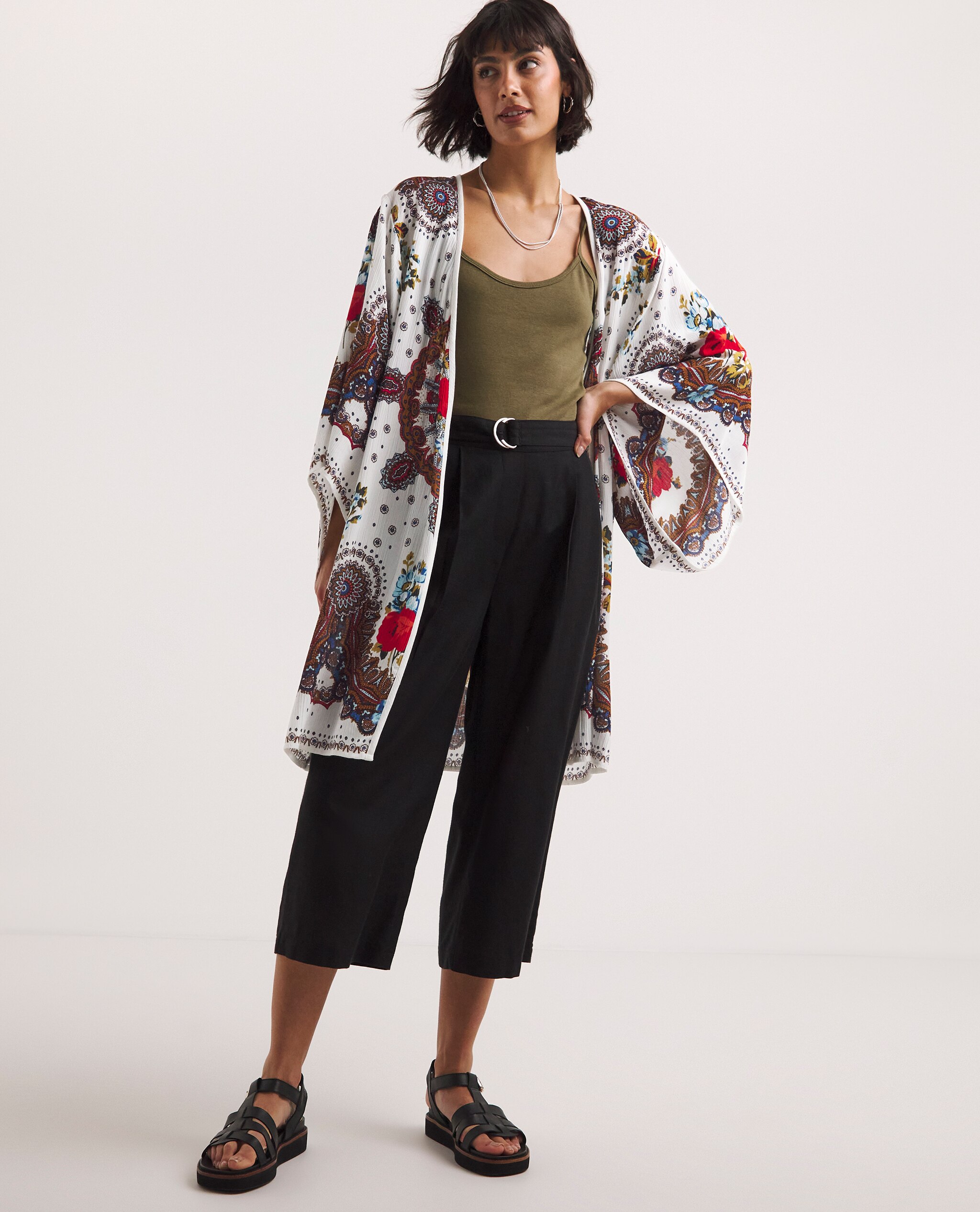 Model is wearing a olive green vest top underneath a print tunic kimono with black cropped trousers and black strap leather sandals.