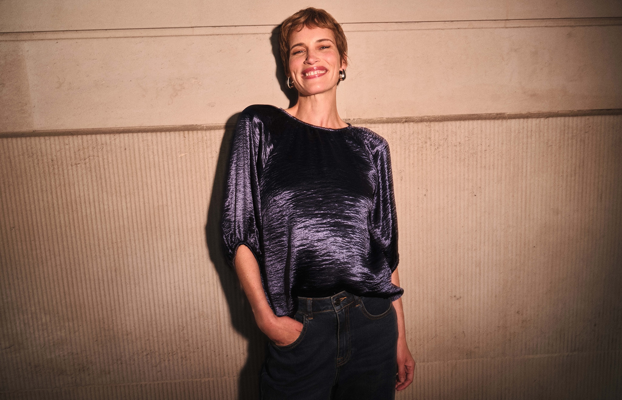 A lady wearing a shiny blue top with dark jeans stood up next to a wall