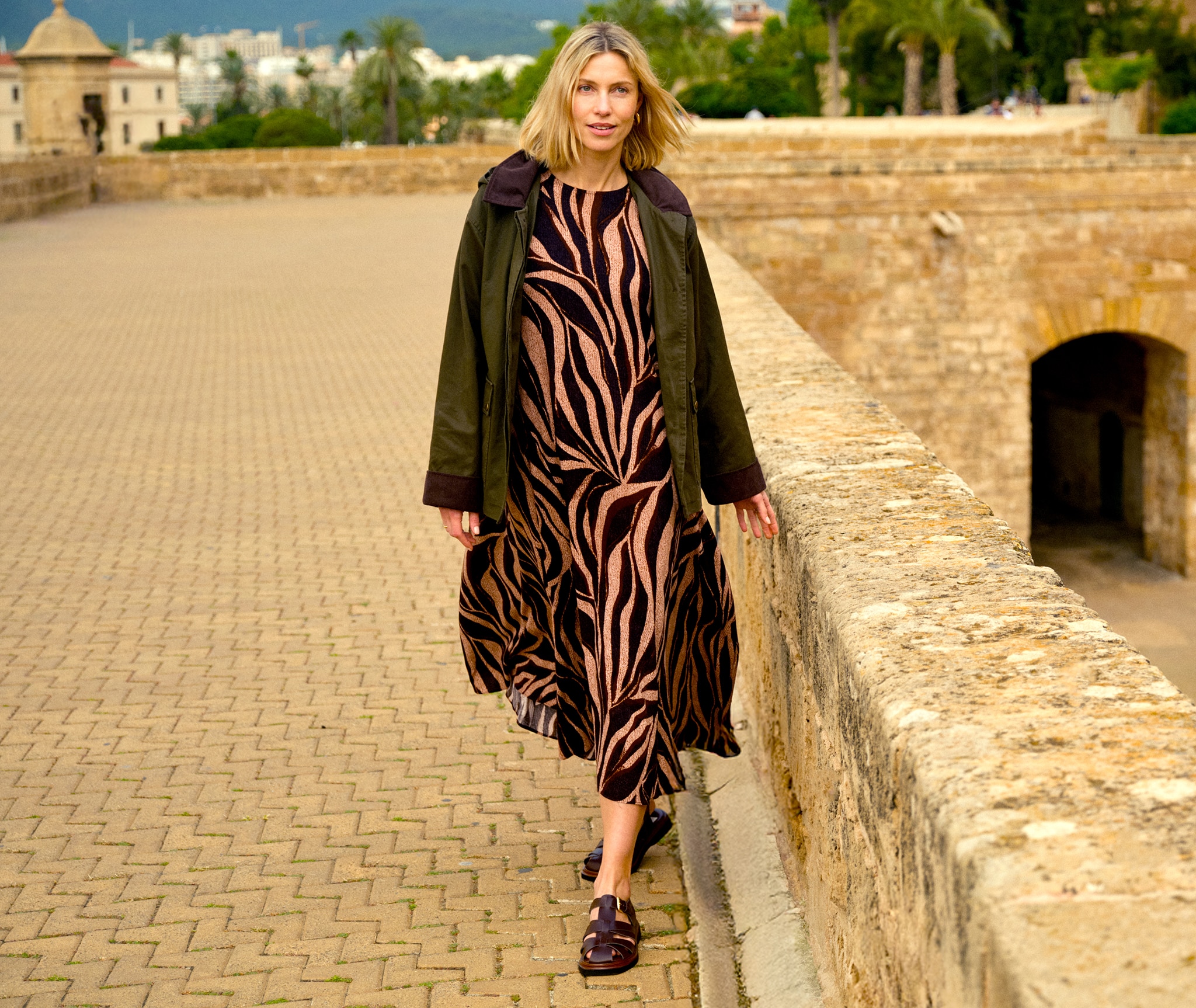 A lady walking next to a stone wall wearing a bronze an black animal print dress.