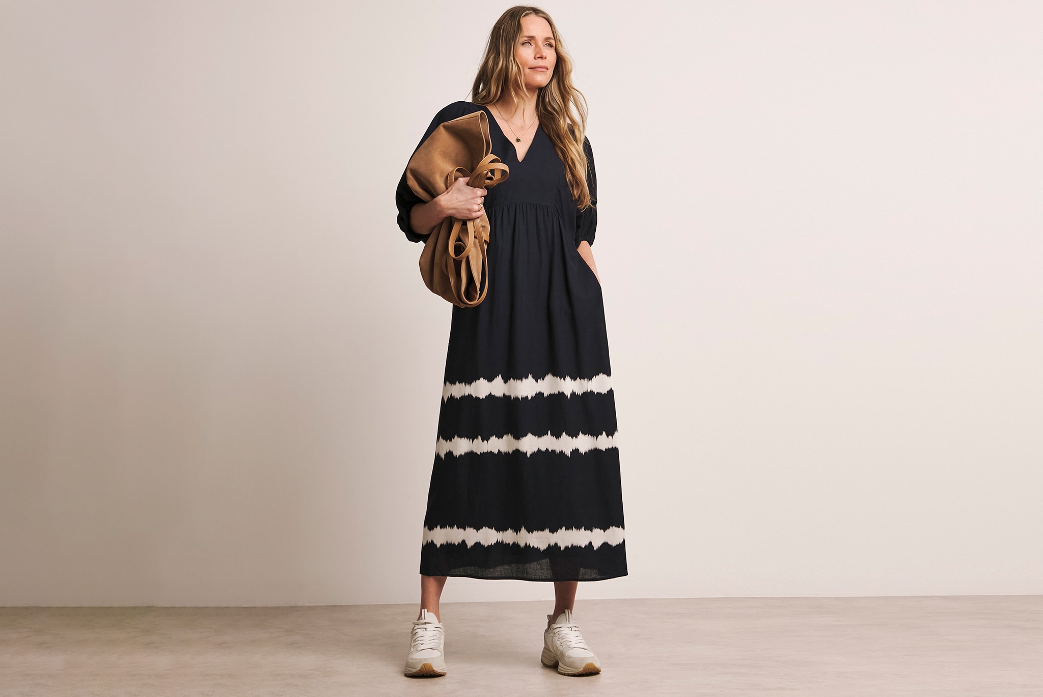 A person wearing a long black dress with white horizontal tie‑dye stripes, paired with white chunky trainers and carrying a large bag.