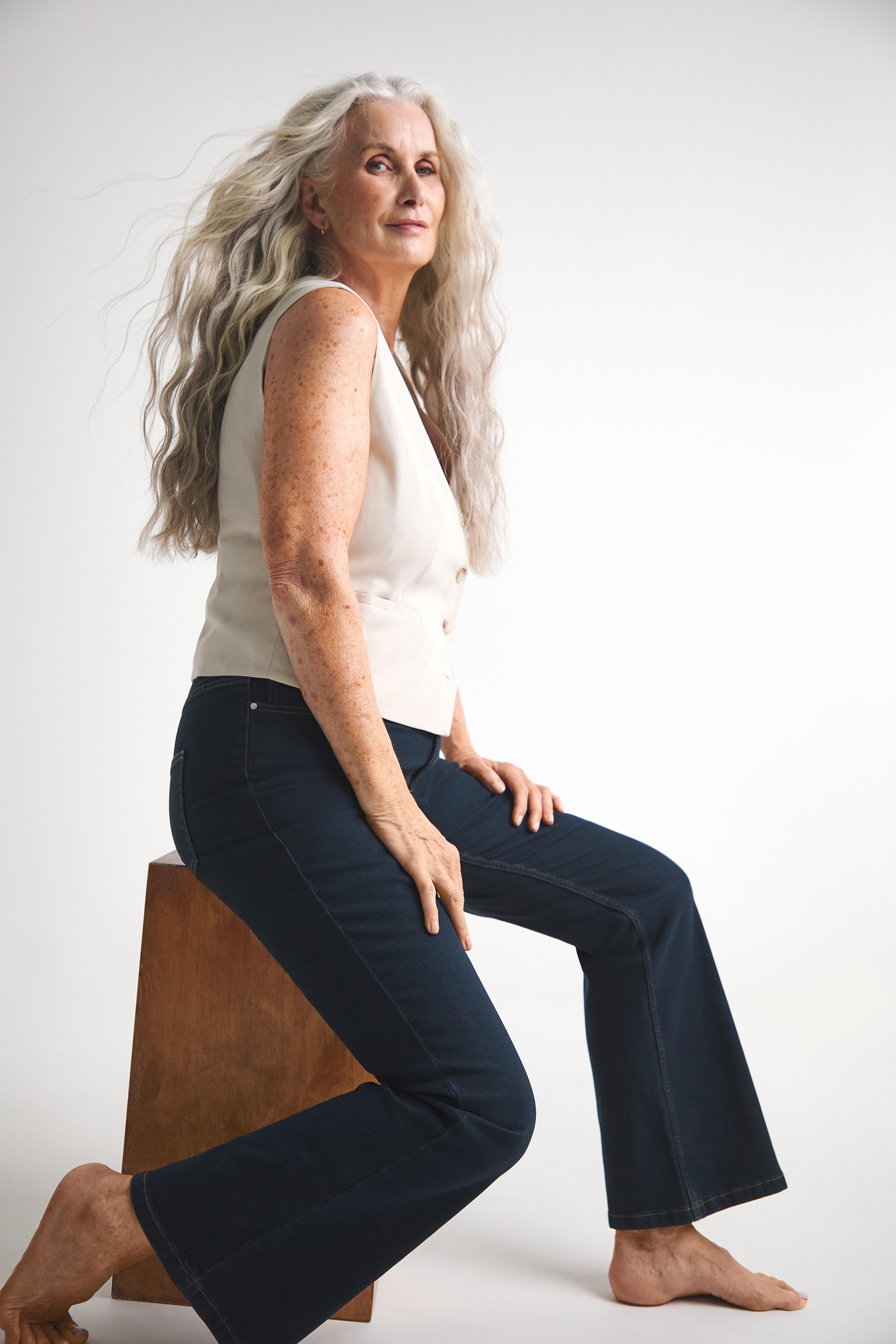 Woman sat on a. box wearing flared dark blue jeans and a cream waistcoat