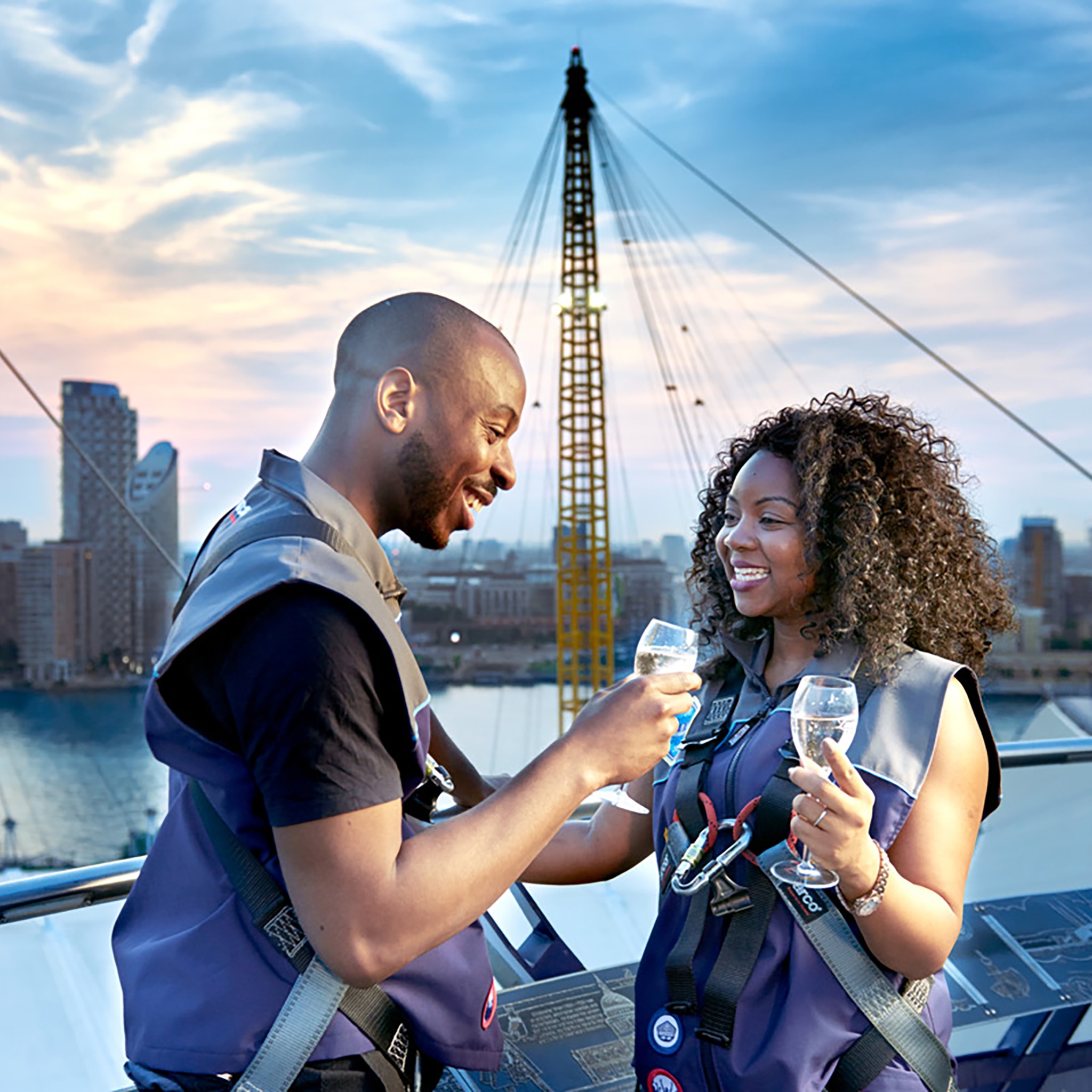 Two people wearing safety harnesses stood on the roof of the 02 arena in London drinking a sparkling drink.