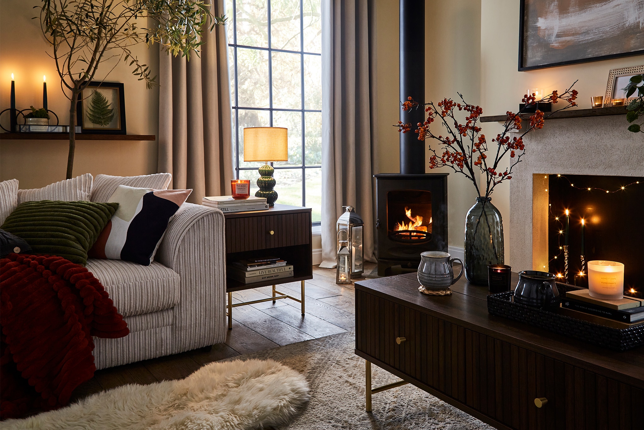 A cosy living room with a sofa, soft lighting, a wood‑burning stove, and candles, decorated with warm-toned cushions, throws, and a coffee table.