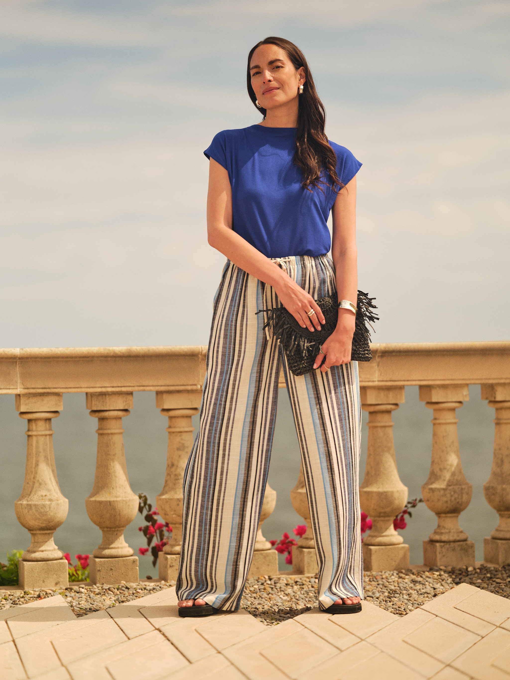 A lady stood next a columned wall by the sea wearing blue striped wide leg trousers.