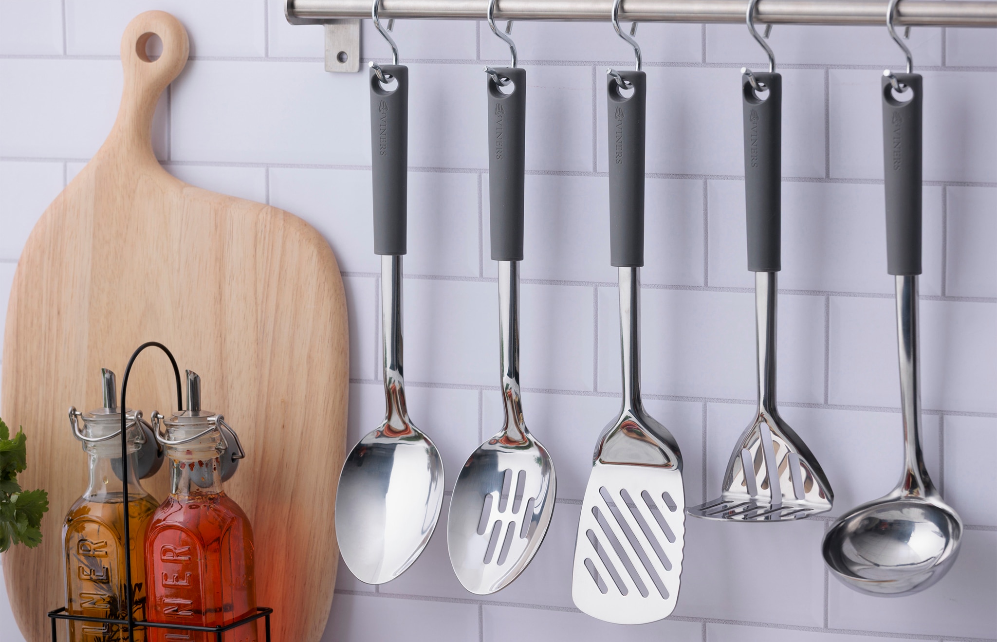 A range of chrome kitchen utensils hung on a wall mounted metal bar.